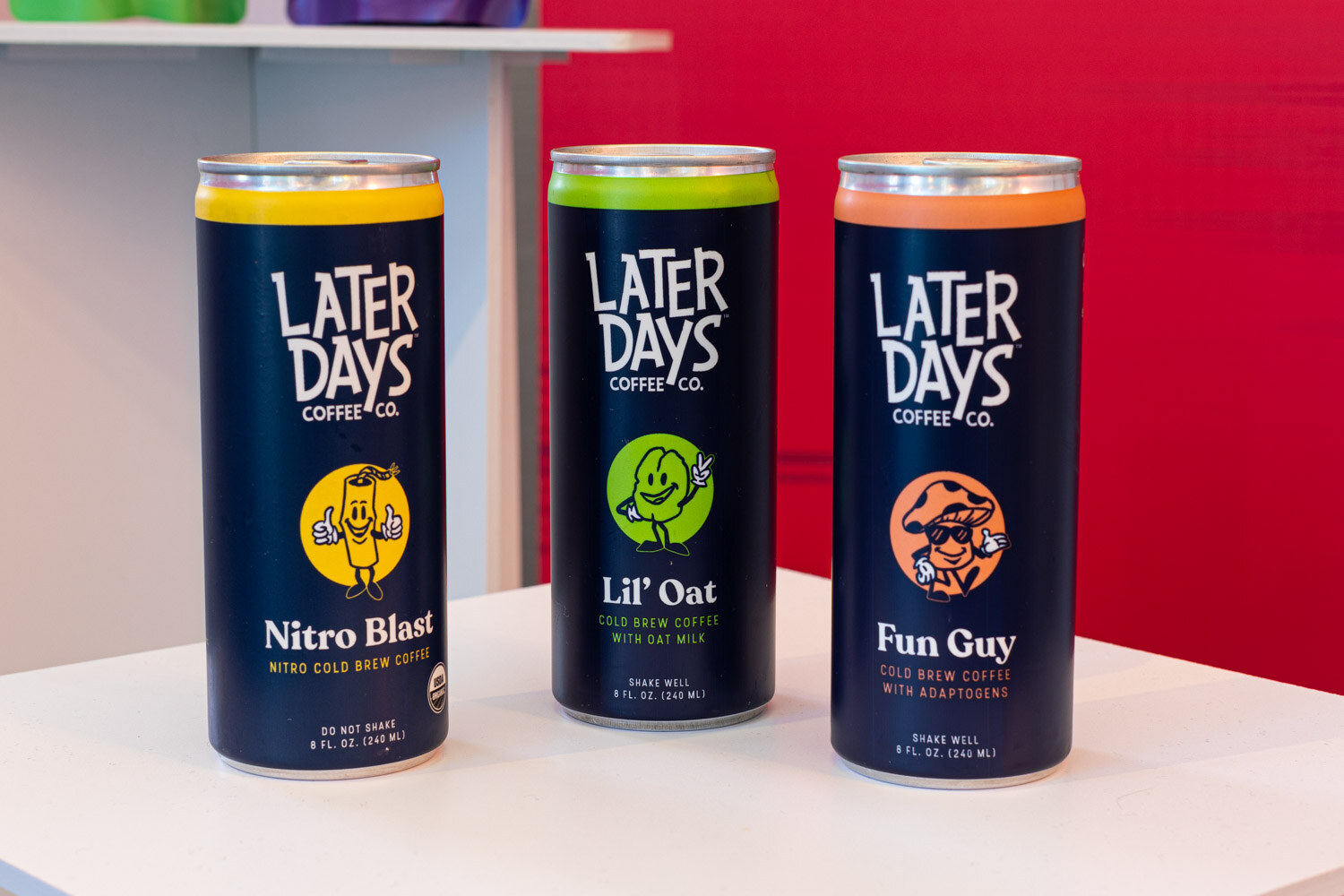 Three navy cans labeled Later Days Coffee Co. Nitro Blast, Lil' Oat and Fun Guy stand on a white table in front of a red wall.