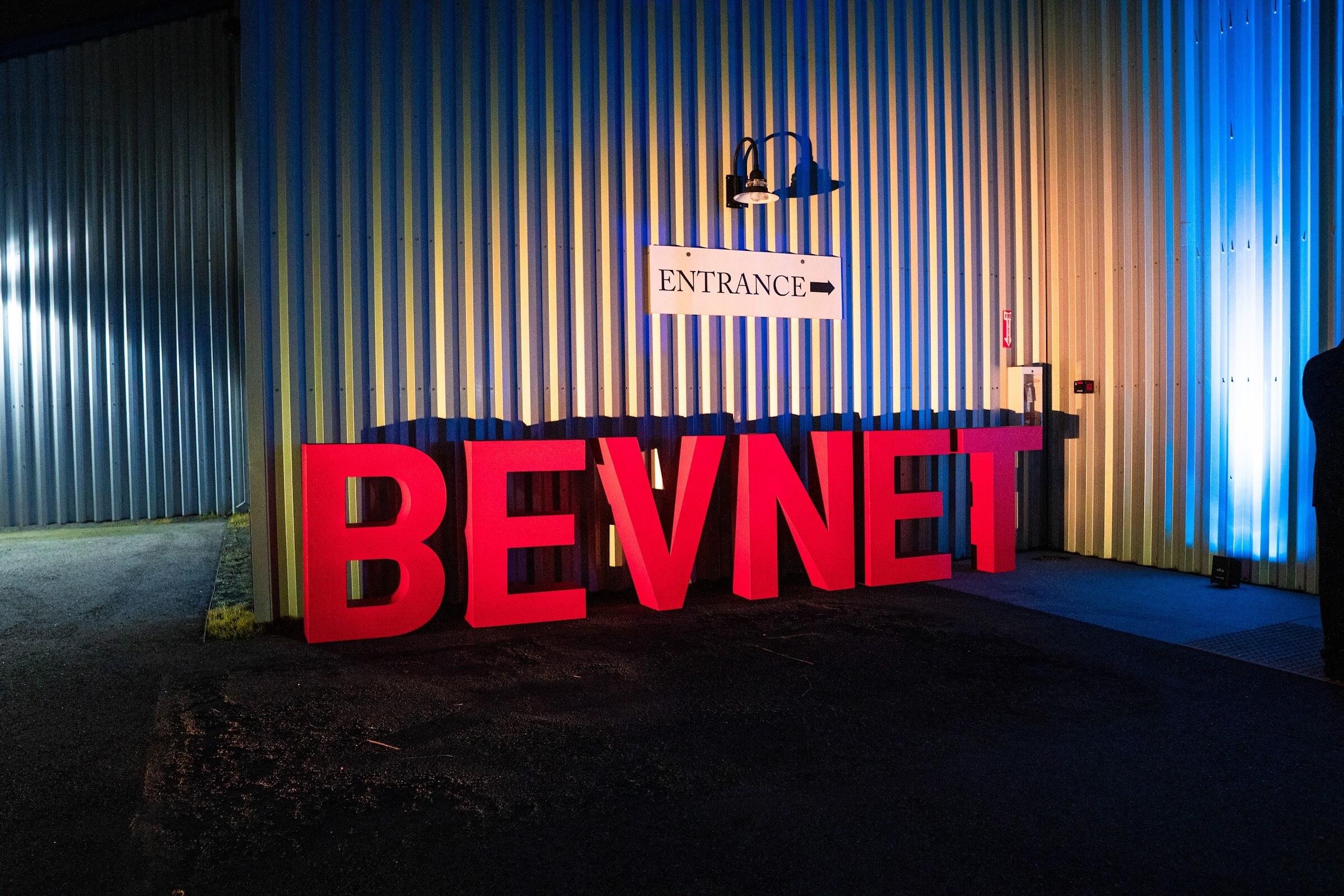 Large red freestanding BEVNET letters in front of a corrugated metal wall lit in blue and gold, with an Entrance sign above.