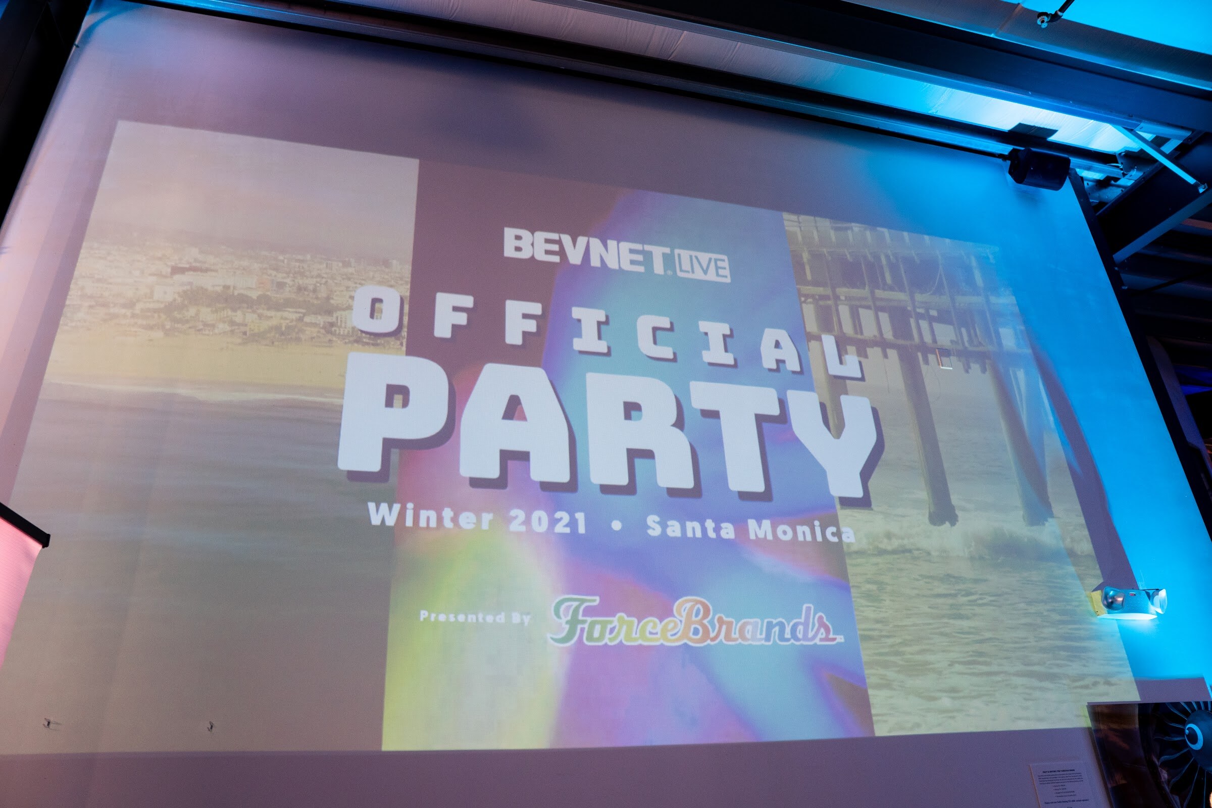 A projection screen at a slight angle reading BevNET Live Official Party Winter 2021 Santa Monica, presented by ForceBrands, over a beach and pier image.