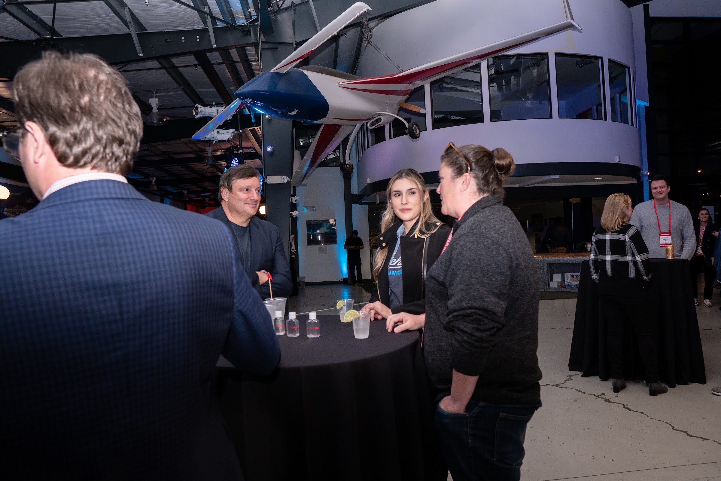 A man in a blazer, a blonde woman and a woman in a gray sweater stand at a black-draped cocktail table with an airplane model hanging overhead.