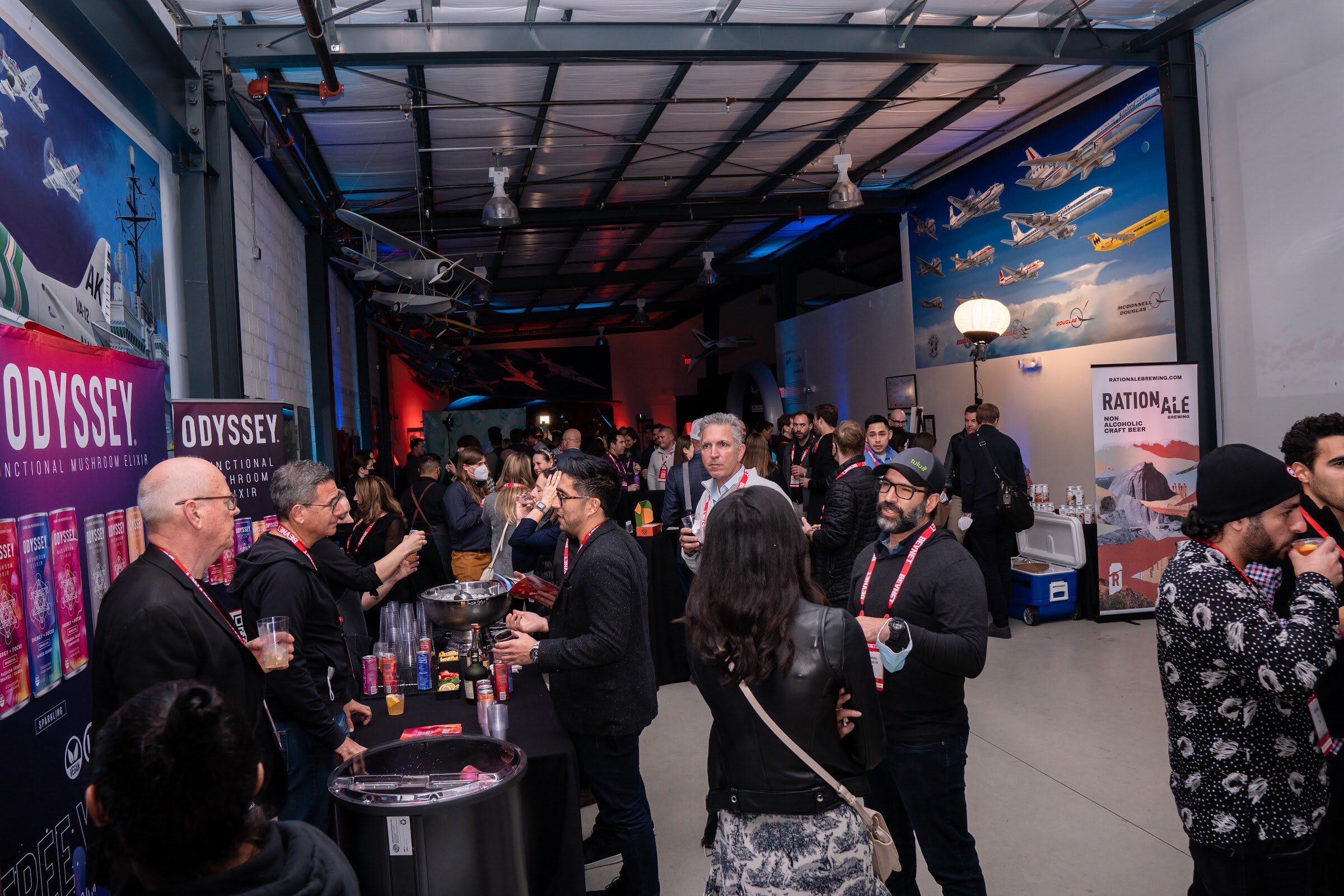 A crowd of attendees on a concrete floor in a hangar with banners for Odyssey and Ration Ale and airplane murals on the walls.