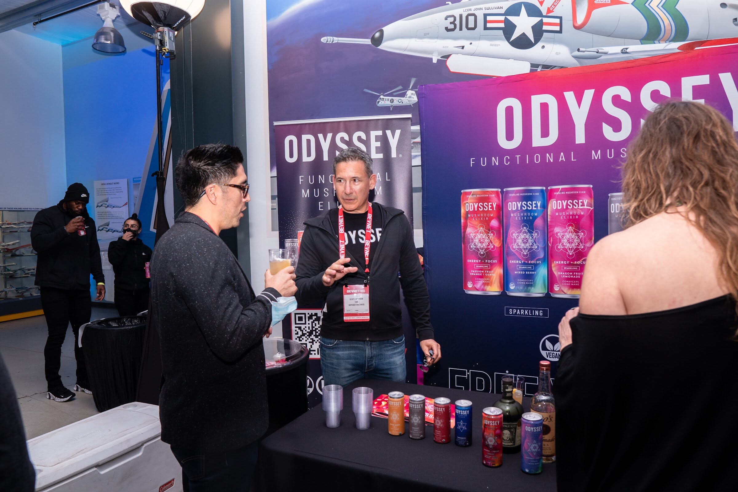 A man in a black hoodie with a BevNET lanyard and a man in a blazer talk in front of a purple Odyssey backdrop with cans lined on a black table.