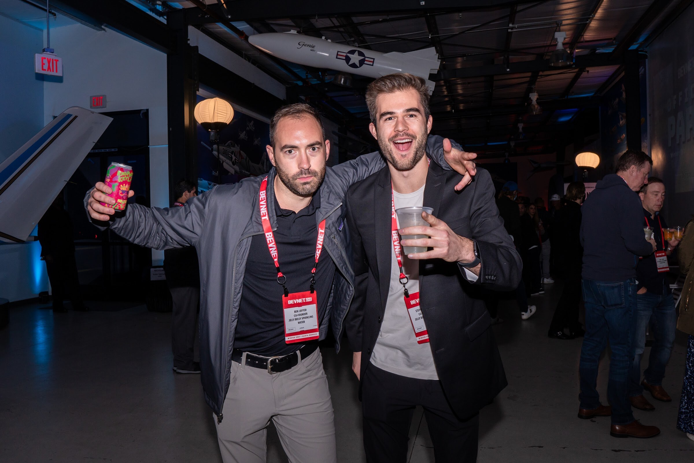 Two smiling men in blazers wearing BevNET lanyards pose close together, one holding a can and one a plastic cup, with a model plane overhead.