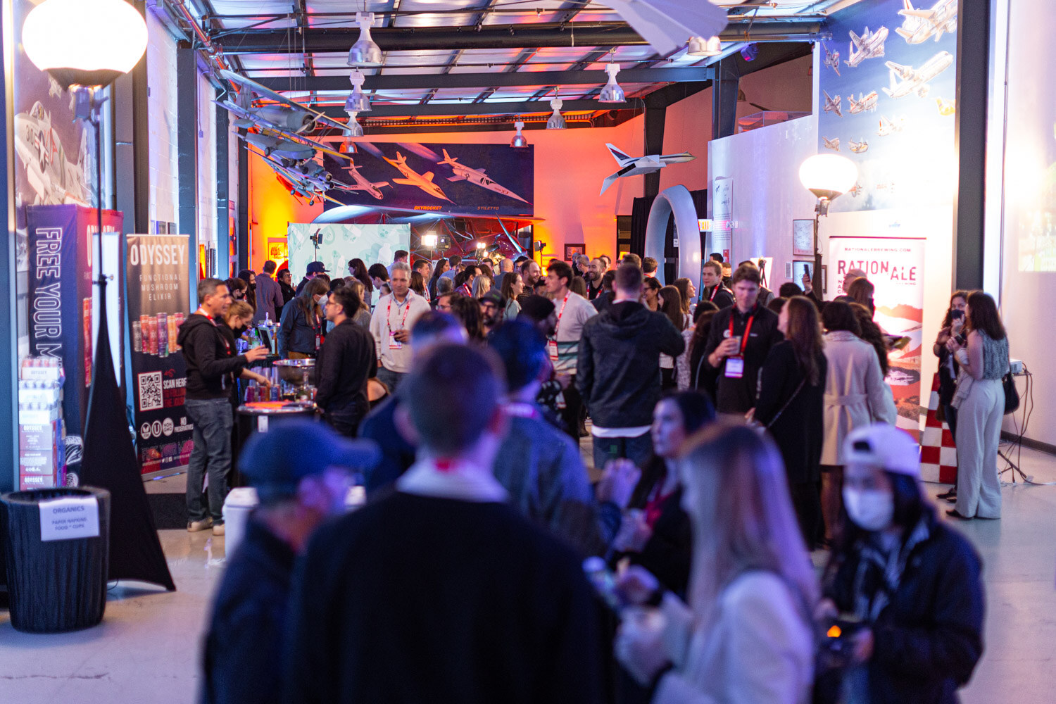A crowd of guests in a long hangar room with orange lighting, aircraft overhead and Odyssey and Ration Ale banners along the wall.