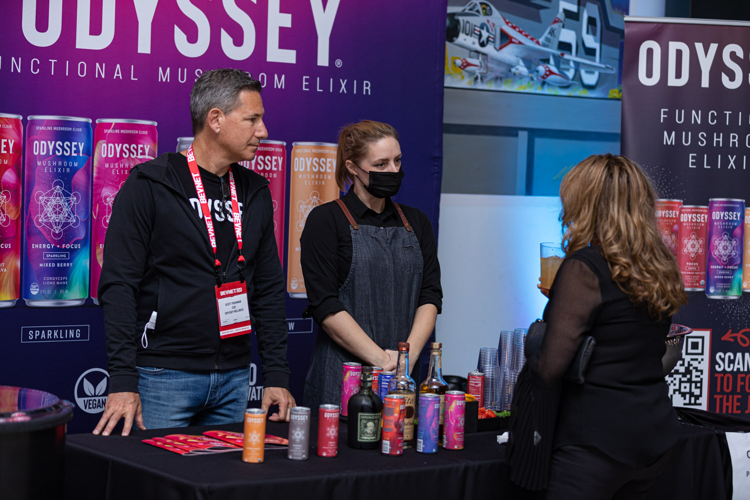 A man in a black Odyssey hoodie and a woman in an apron stand behind a black table with Odyssey cans and bottles in front of a purple Odyssey backdrop.