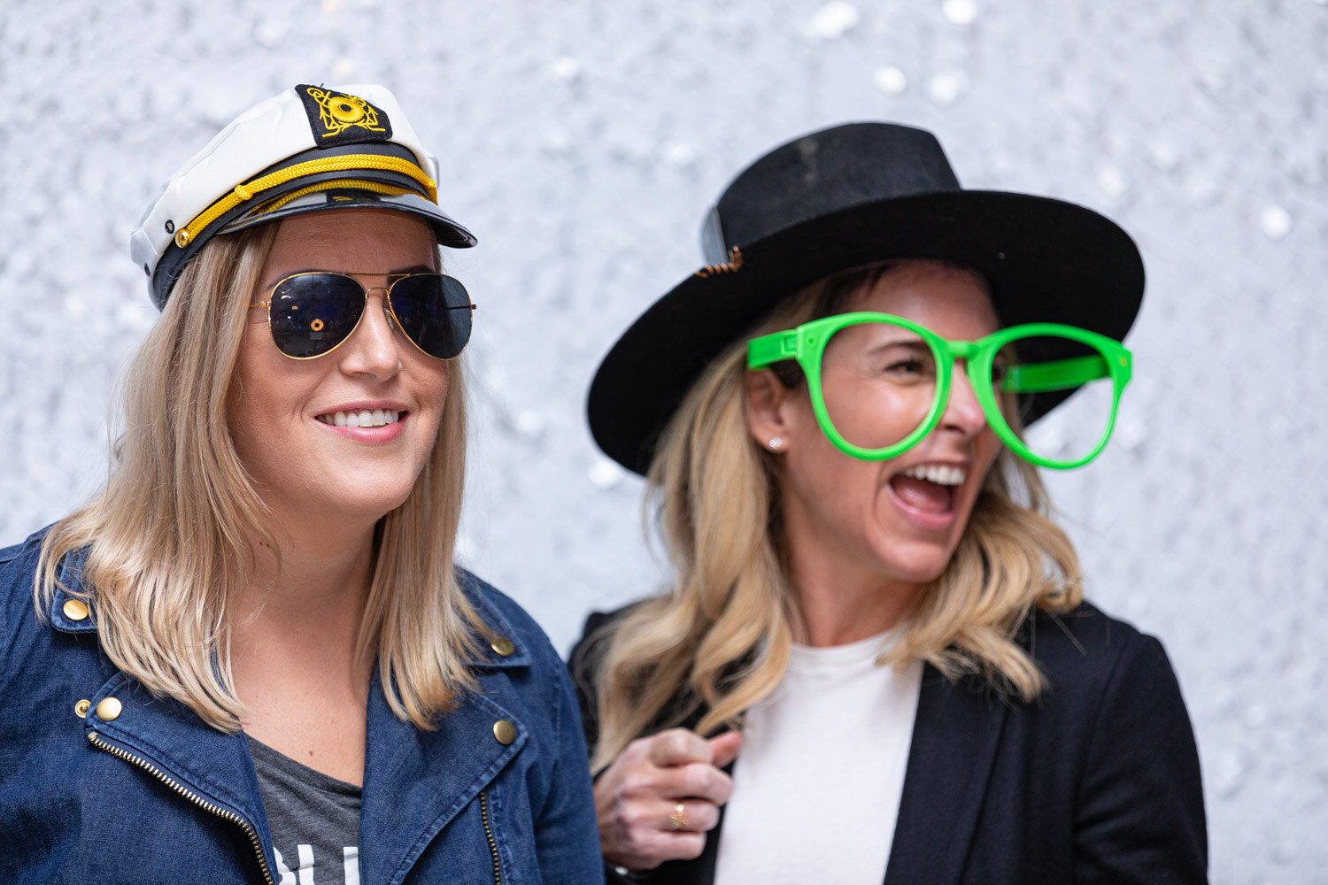 A woman in a white captain's hat and aviator sunglasses stands beside a laughing woman in a black hat and large green glasses in front of a sparkling backdrop.