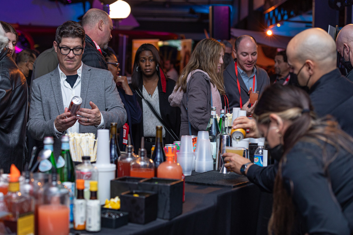 A man in a gray blazer holds a can behind a black-draped bar covered with bottles, mixers and cups while a bartender mixes a drink.