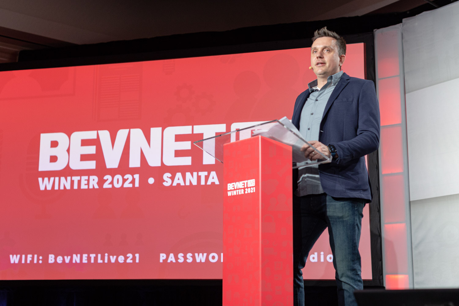 A man in a navy blazer speaks at a red podium in front of a red screen reading BevNET Winter 2021 Santa Monica with Wi-Fi details.