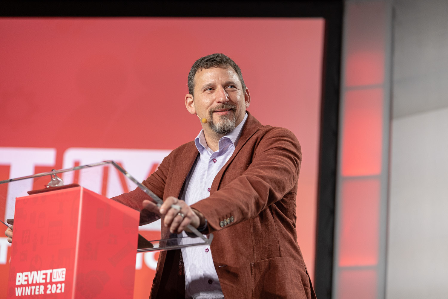 A bearded man in a rust-colored jacket stands at a clear lectern with a red BevNET Live Winter 2021 podium in front of a red backdrop.