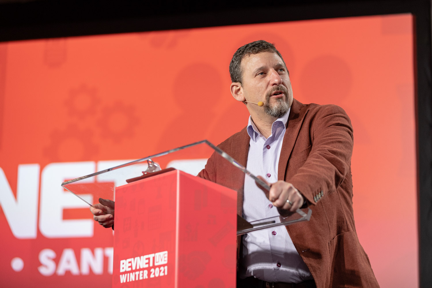 A bearded man in a rust jacket grips a clear lectern in front of a red BevNET Live Winter 2021 podium and a red screen.