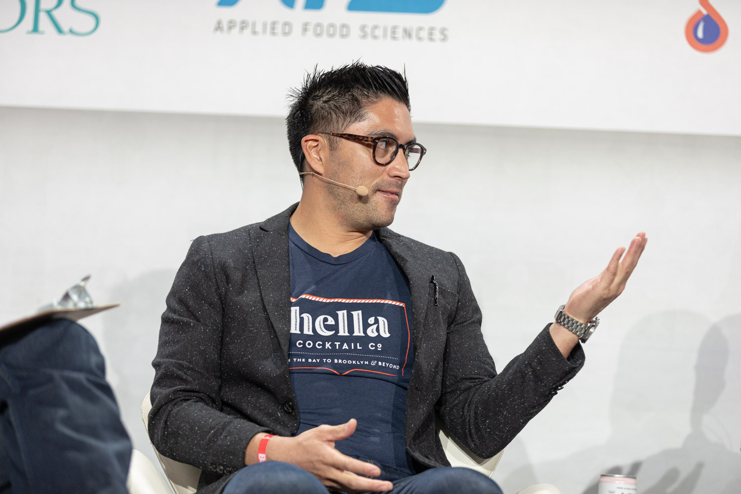 A man with glasses and a navy Hella Cocktail Co. t-shirt under a gray blazer gestures with an open hand while seated on stage.