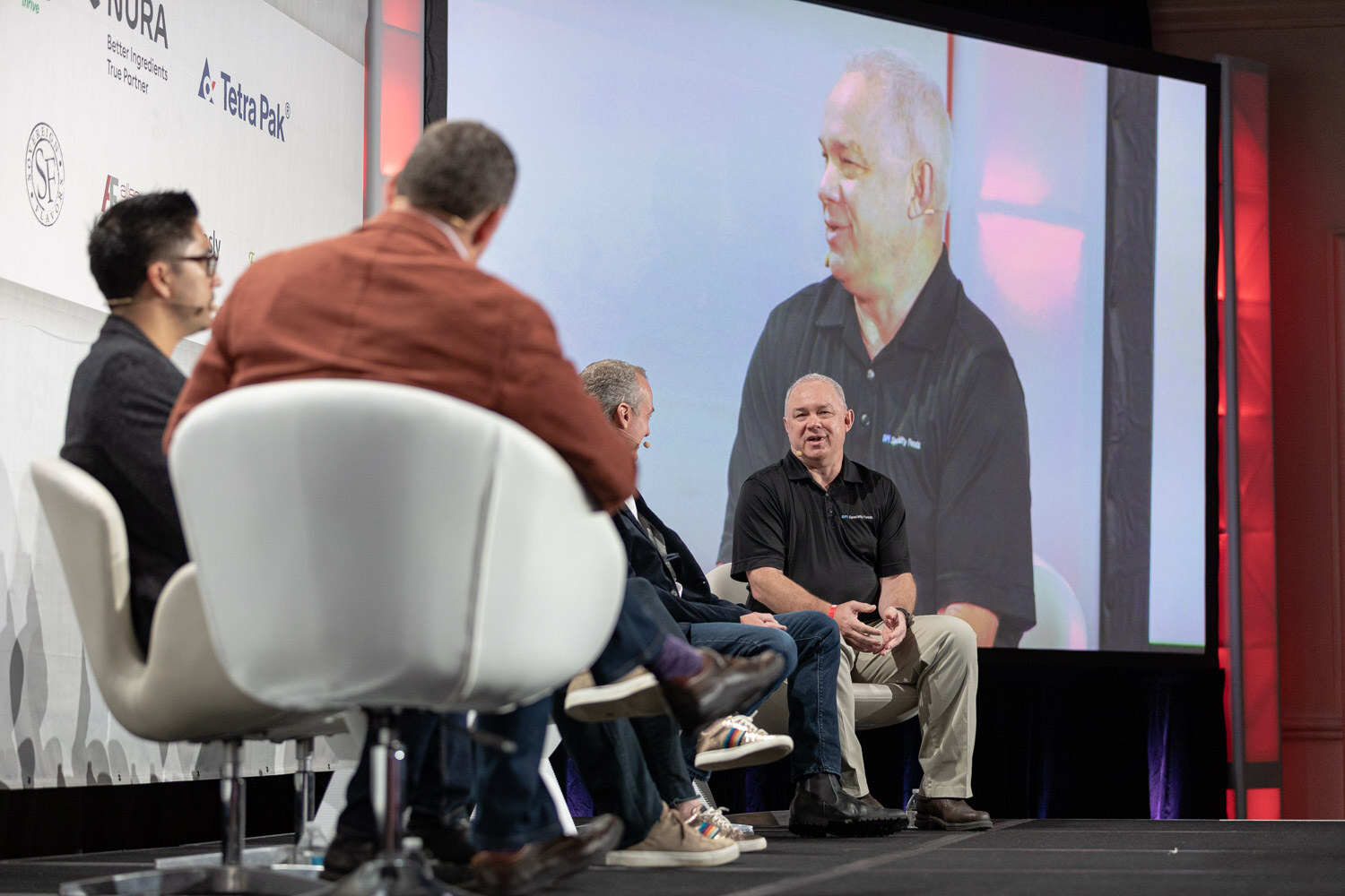 A view over the shoulder of a man in a rust jacket toward four panelists seated in white chairs, with a screen showing a smiling man in a black polo.