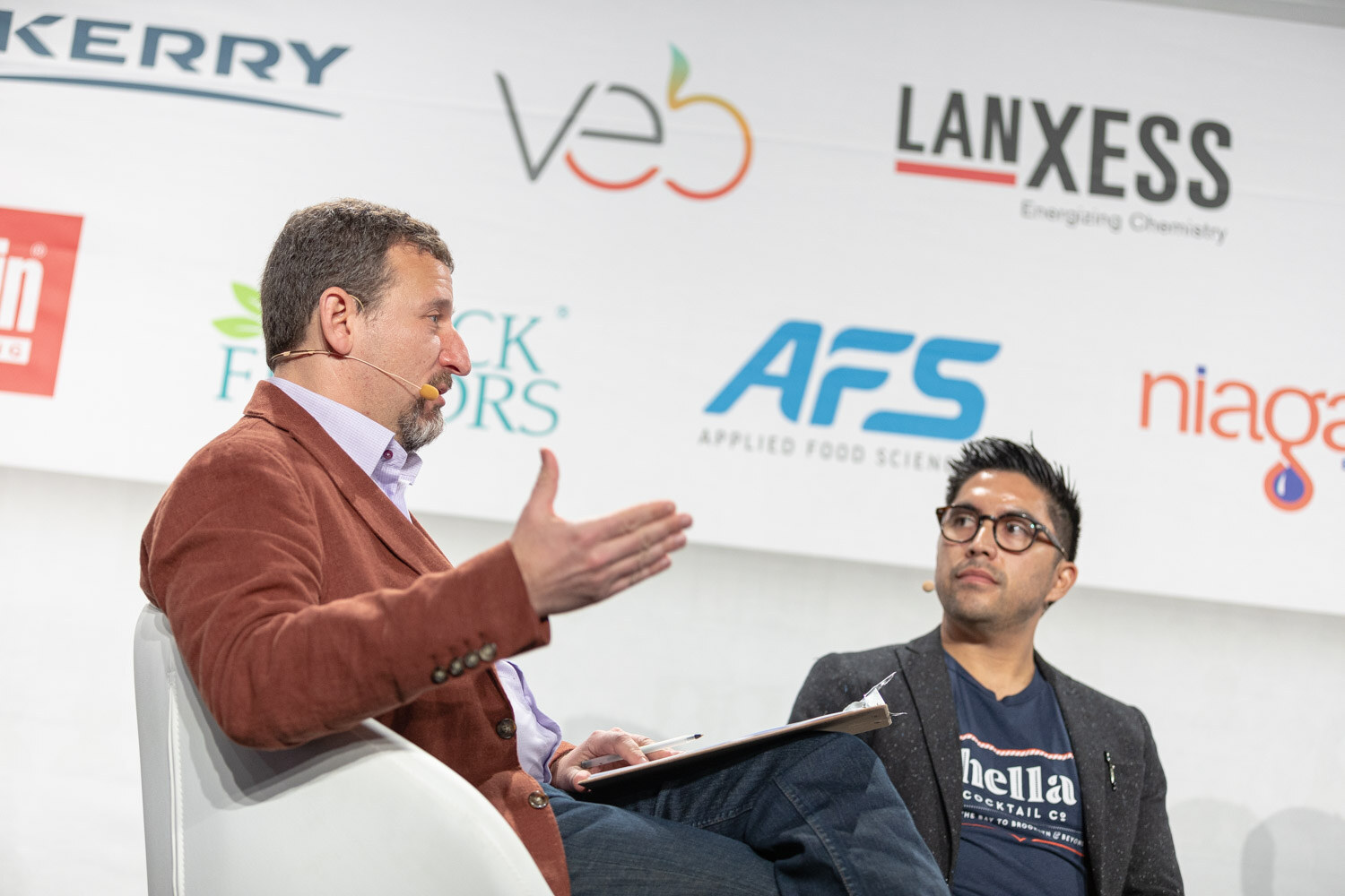 Two men seated in white chairs in front of a sponsor wall, one in a rust jacket gesturing and one in a navy Hella Cocktail Co. shirt looking at him.