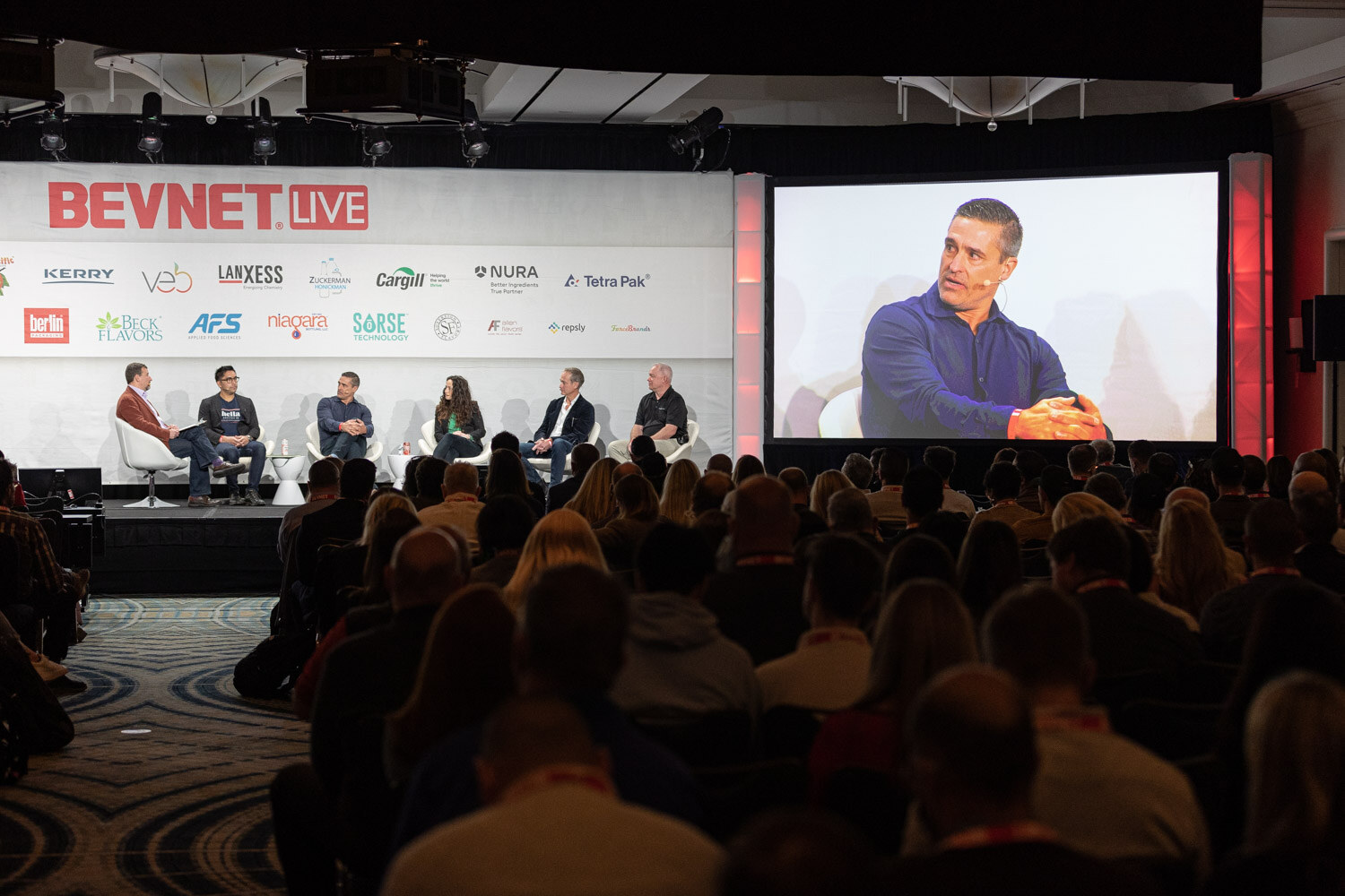 A wide view of the BevNET Live stage with six people seated in white chairs, a large screen showing a man in a blue sweater, and a seated audience in the foreground.
