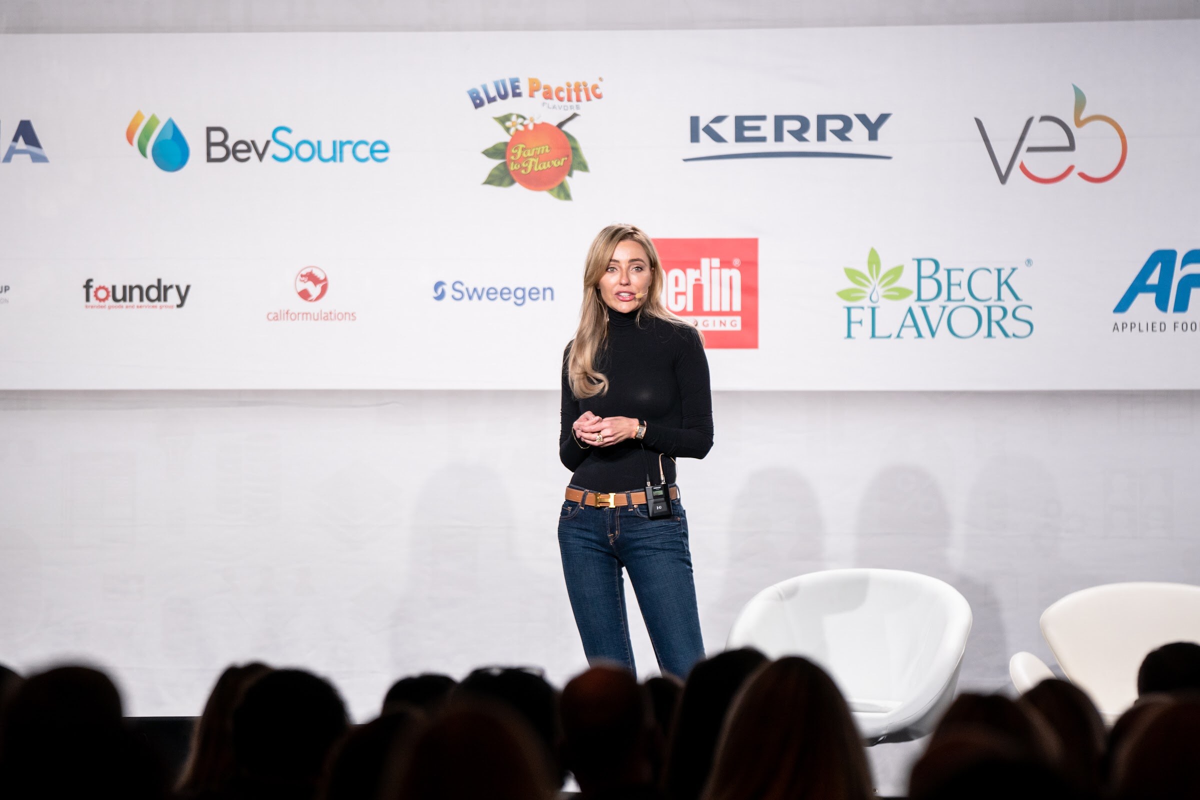 A woman with long blonde hair in a black top and jeans stands on stage in front of a sponsor wall with BevSource, Kerry, veb, Berlin Packaging and Beck Flavors logos.