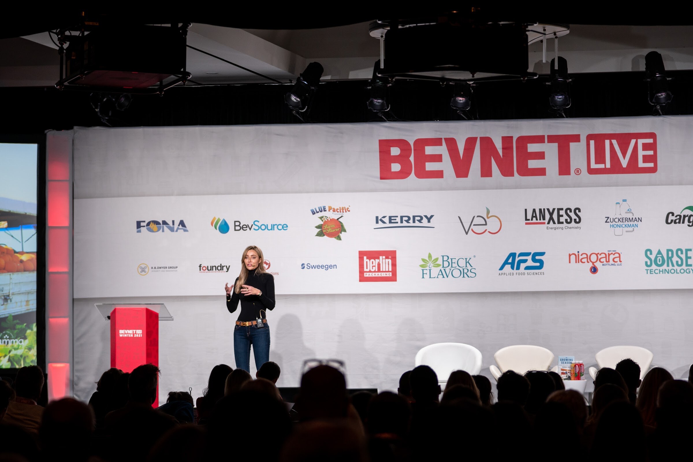 A woman in black stands on a large stage beneath a BevNET Live banner and sponsor logos, with a red podium to her left and audience heads in silhouette.