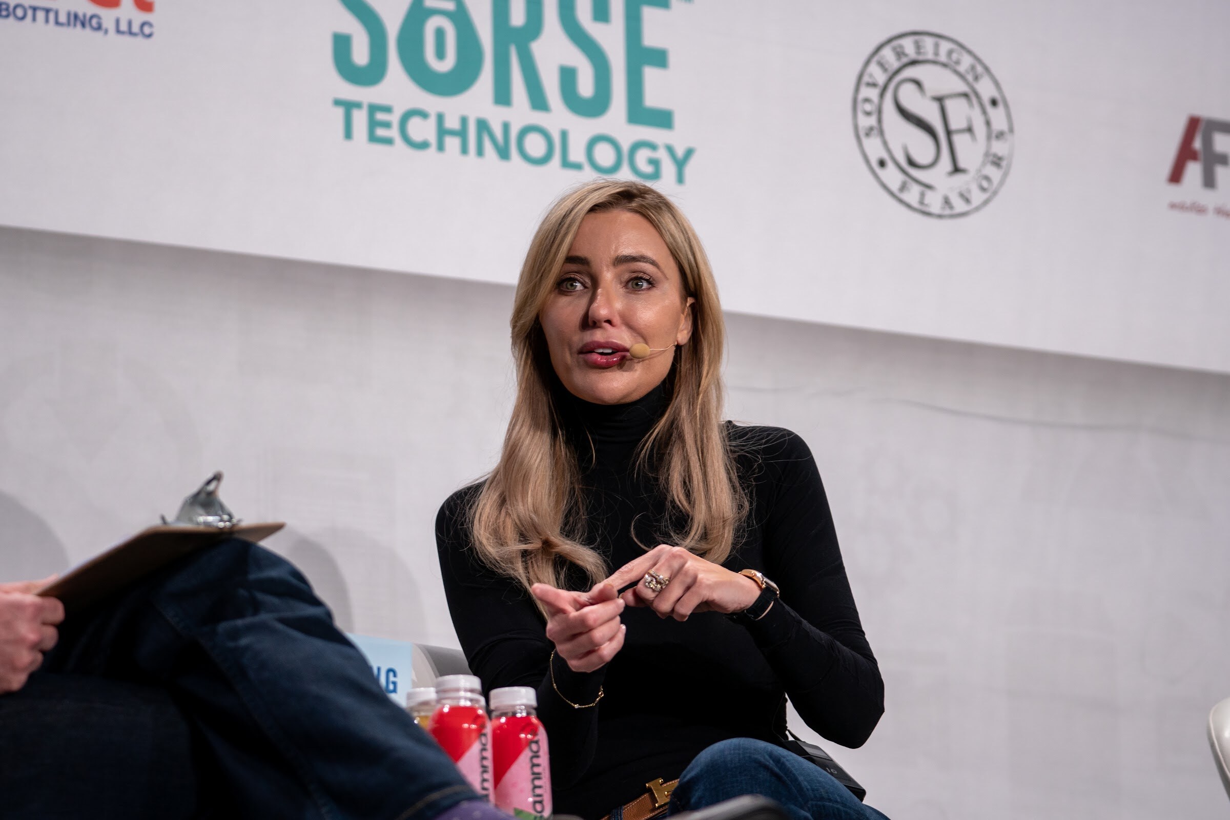 A blonde woman in a black sweater sits in a white chair, hands clasped, with red bottles at the lower edge and a Sorse Technology sign behind her.