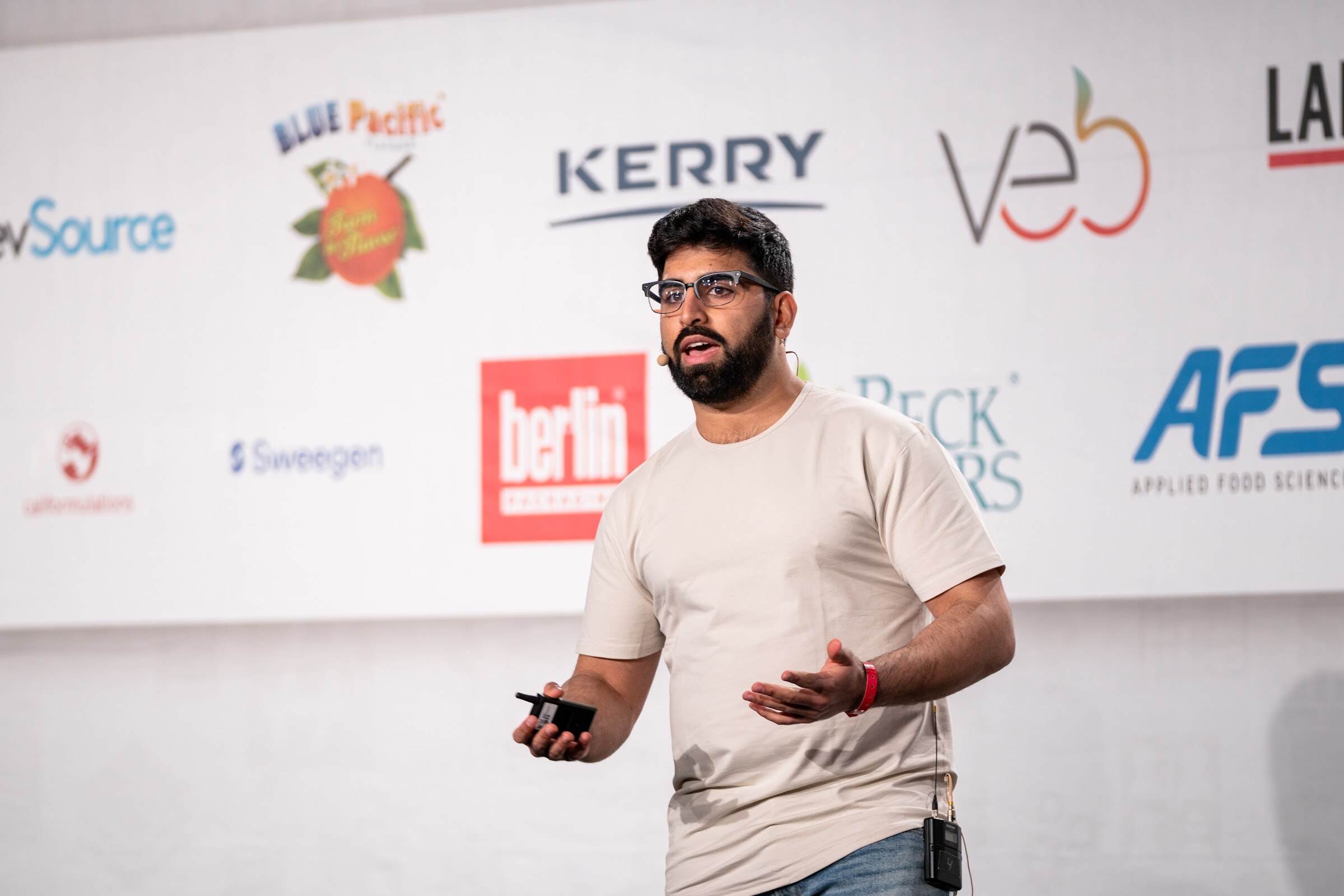 A bearded man in glasses and a cream t-shirt gestures with a clicker in his hand in front of a sponsor wall including Kerry, veb, Berlin Packaging and AFS.