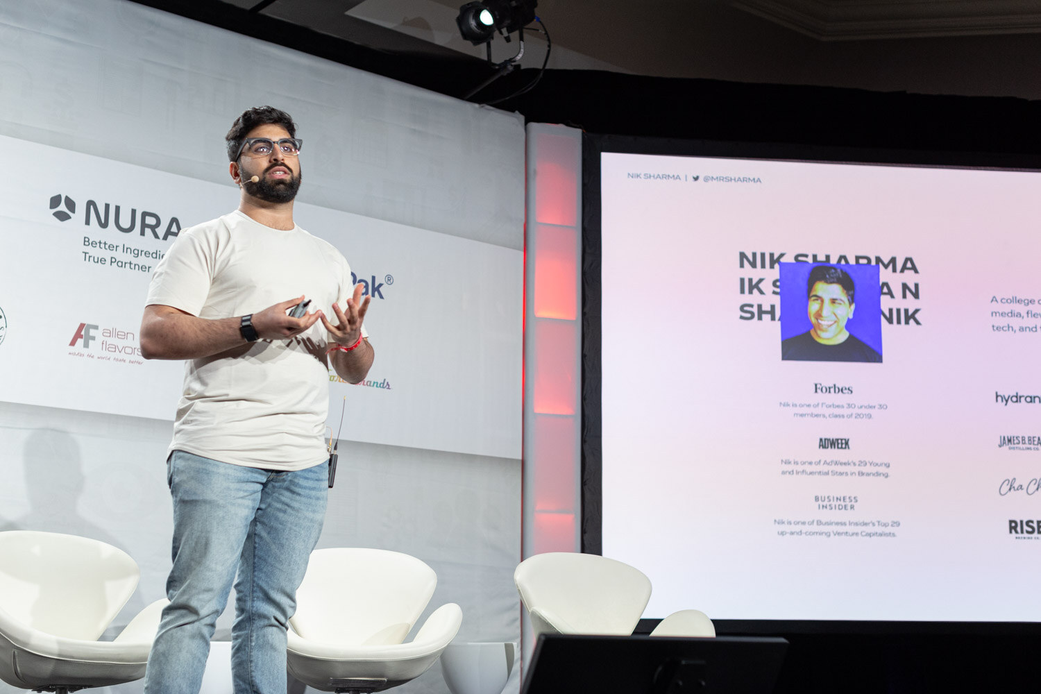 A man in a cream t-shirt stands on stage next to a large slide showing a photo and a list of press mentions under the name Nik Sharma.