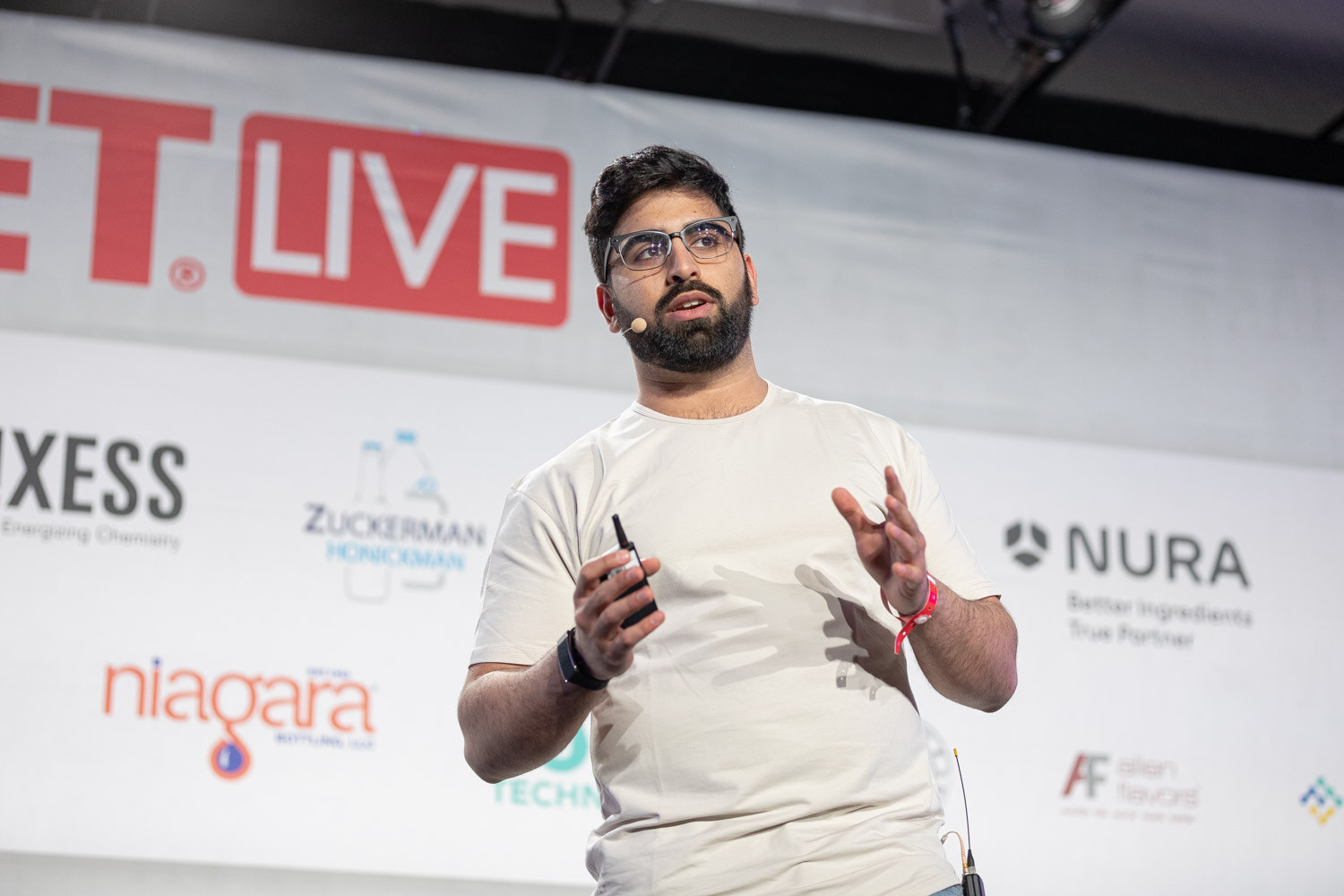 A bearded man in glasses and a cream t-shirt gestures with both hands in front of a sponsor wall with Lanxess, Zuckerman Honickman, Nura and Niagara logos.