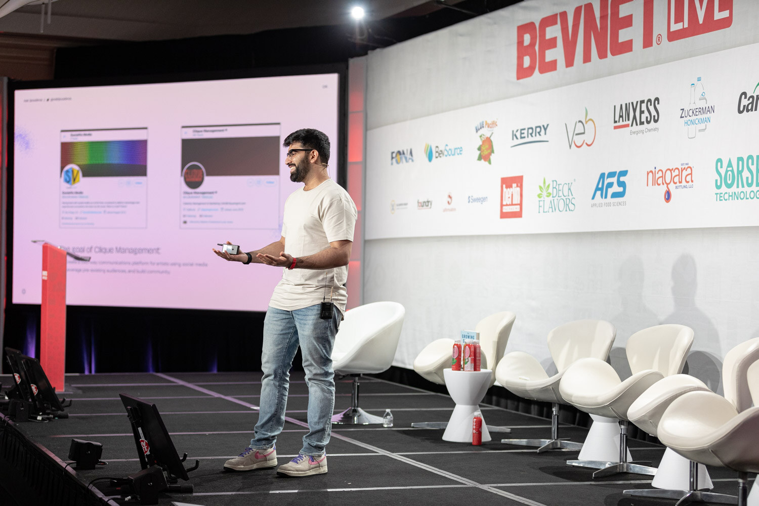 A man in a cream t-shirt and jeans stands on a long stage beside empty white chairs while a large screen behind him shows two social media profile pages.