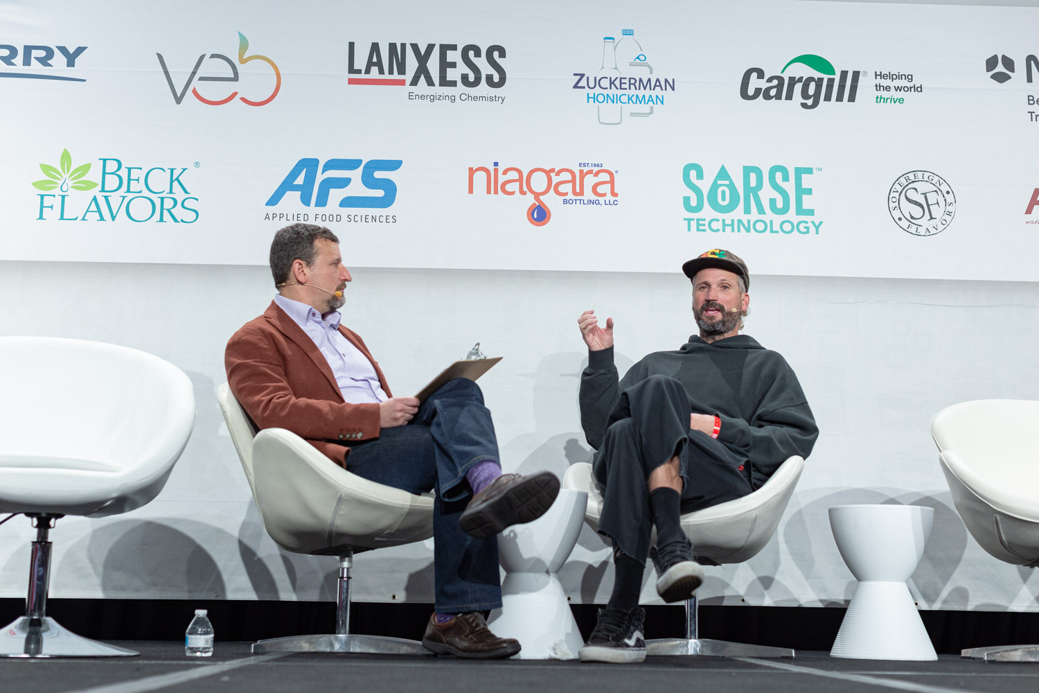 A man in a rust jacket with a clipboard and a man in a black hoodie and colorful cap sit in white chairs in front of a sponsor wall.