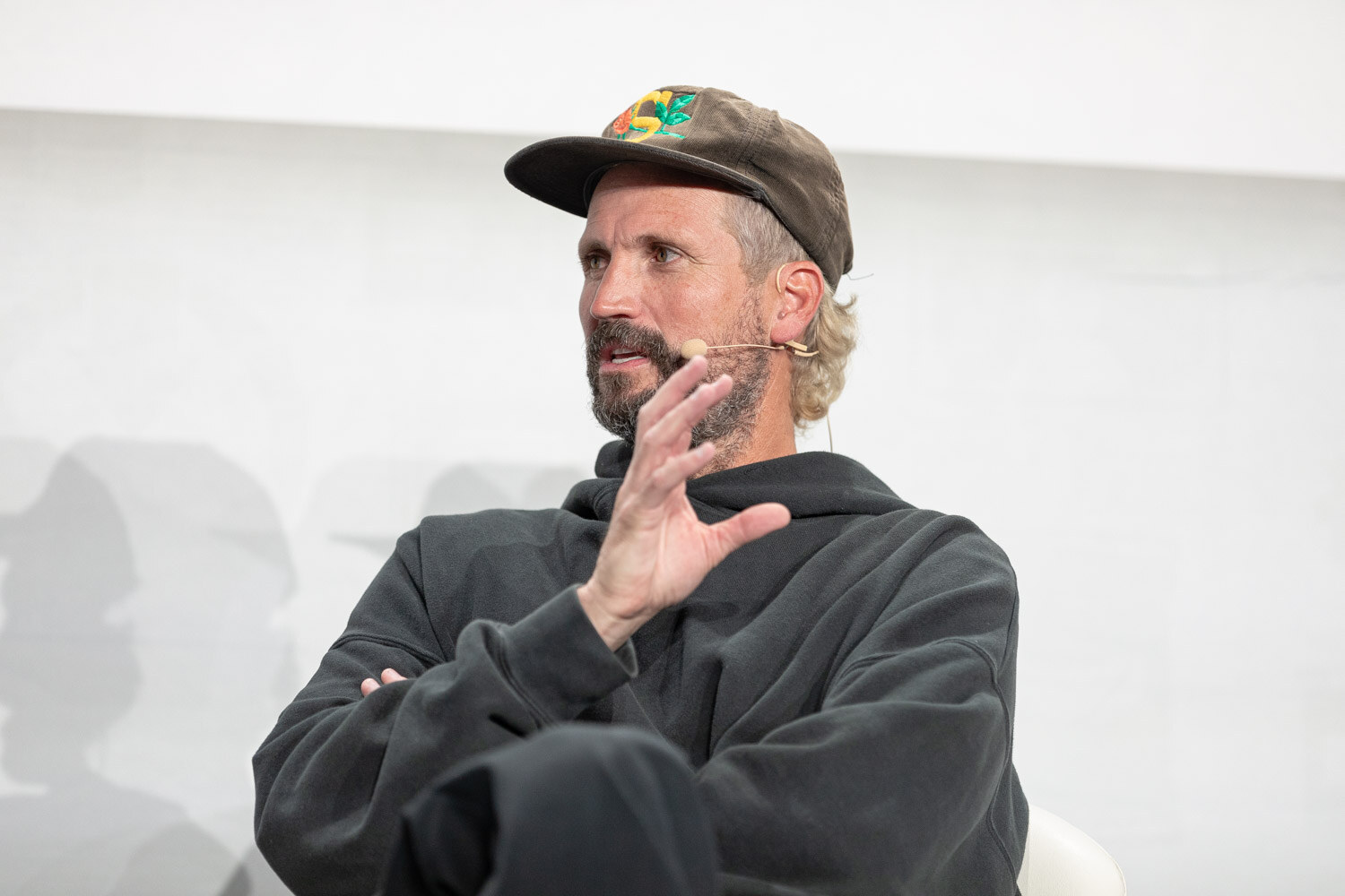 A bearded man in a camouflage cap and black hoodie with a headset microphone raises one hand while looking off to the side.