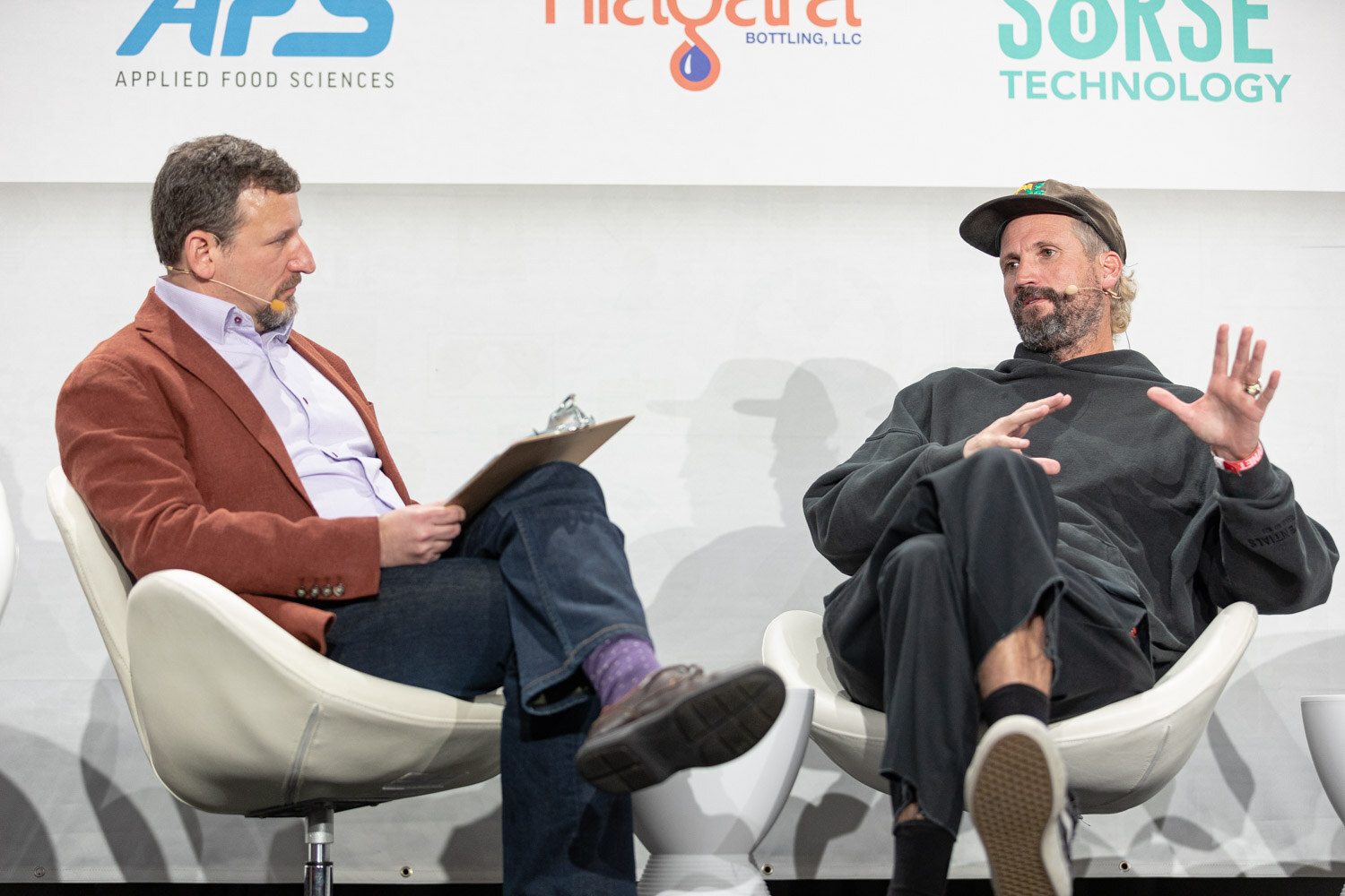 Two men seated in white chairs on stage, one in a rust jacket with a clipboard and one in a dark hoodie and cap raising both hands as he talks.