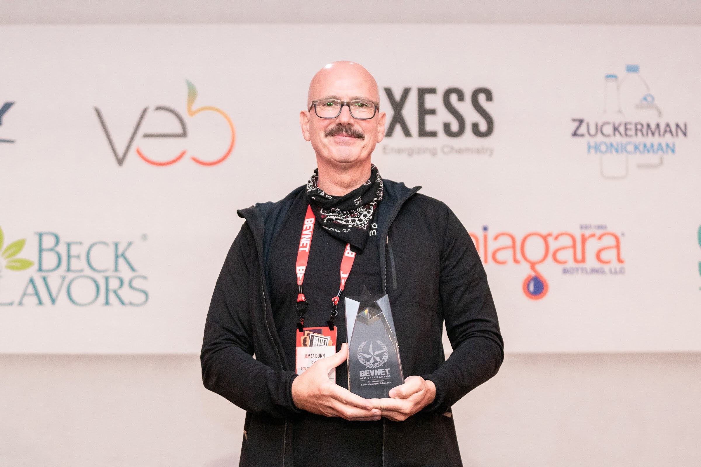 A bald man with glasses and a mustache holds a small glass trophy in both hands in front of a sponsor wall with veb, Beck Flavors and Niagara logos.