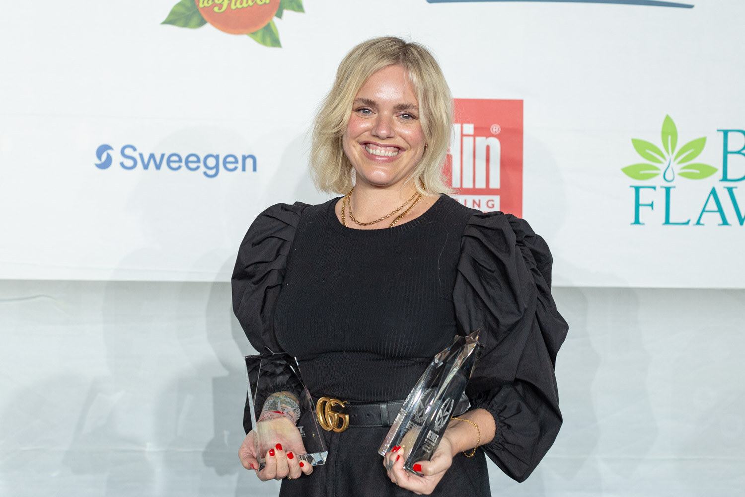 A blonde woman in a black ruffled top holds a glass award in each hand in front of a sponsor wall with Sweegen and Berlin Packaging logos.