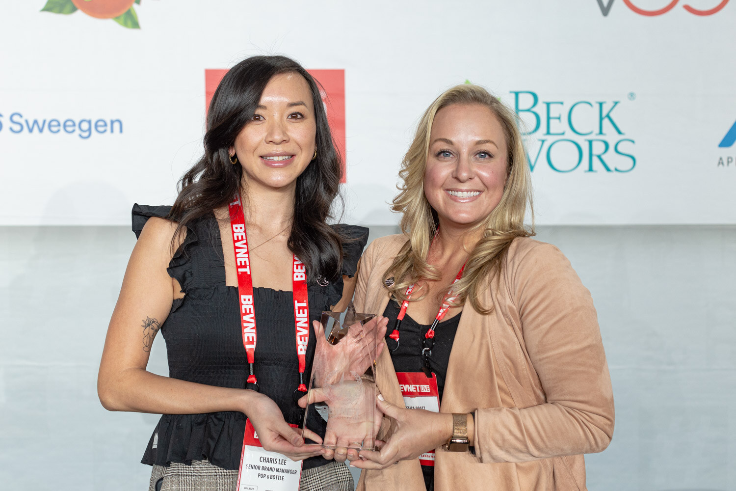 Two smiling women wearing BevNET lanyards stand together holding a glass trophy in front of a sponsor wall with Sweegen and Beck Flavors logos.