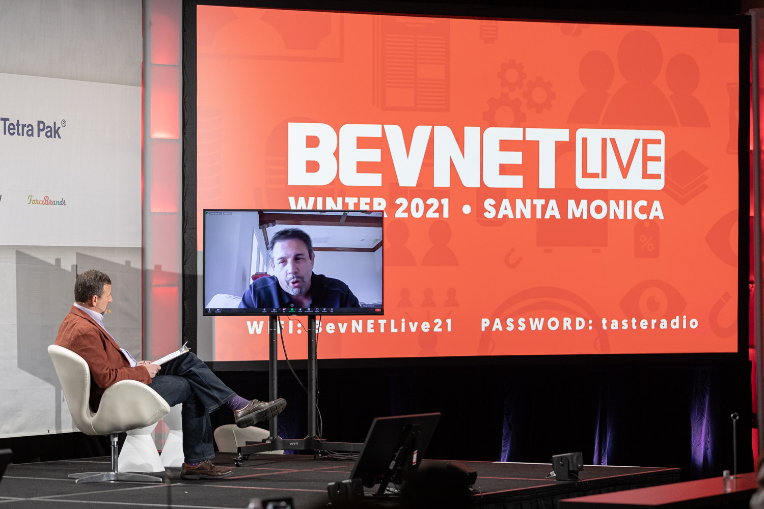 A man in a rust jacket sits in a white chair at the left of the stage while a small video screen shows a remote guest in front of a red BevNET Live Winter 2021 Santa Monica screen.