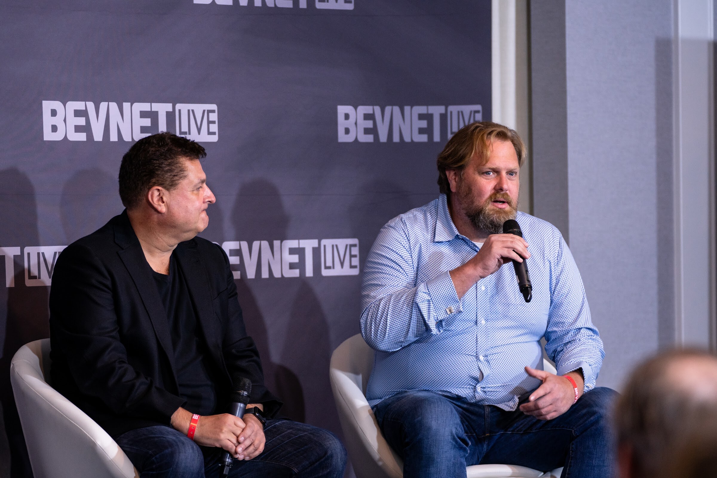 A man in a black jacket and a bearded man in a blue checked shirt holding a microphone sit in white chairs in front of a purple BevNET Live wall.