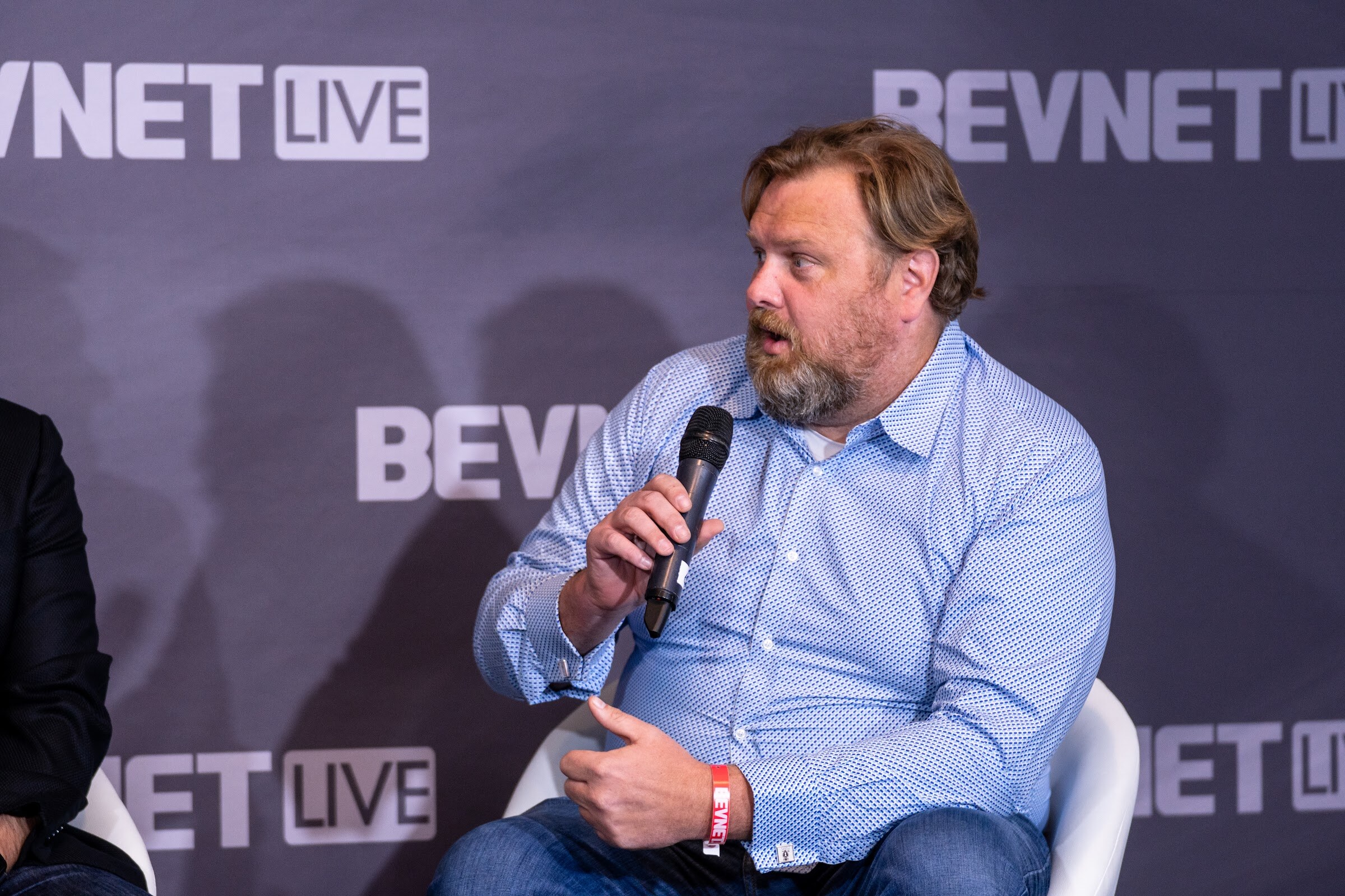 A bearded man in a light blue shirt holds a microphone and looks off to the side in front of a purple BevNET Live backdrop.