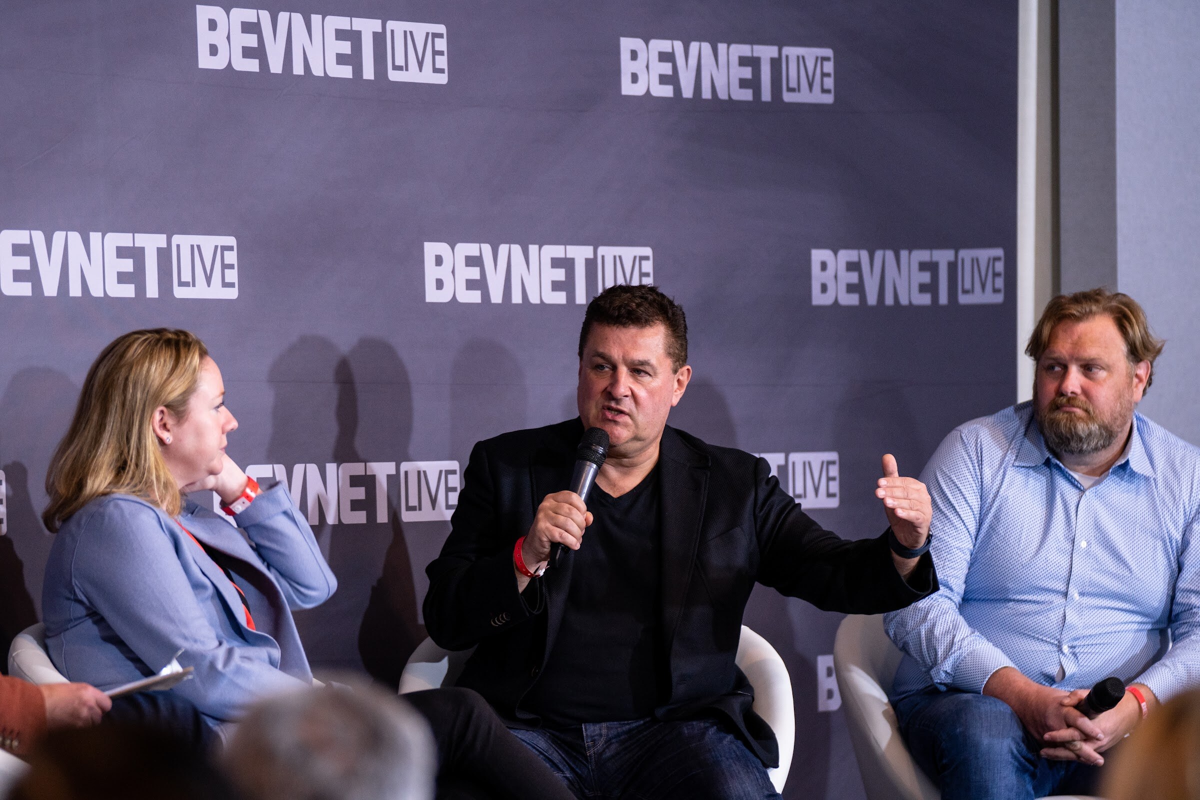 Three people seated in front of a BevNET Live backdrop, with a man in a black jacket in the middle speaking into a microphone and gesturing.