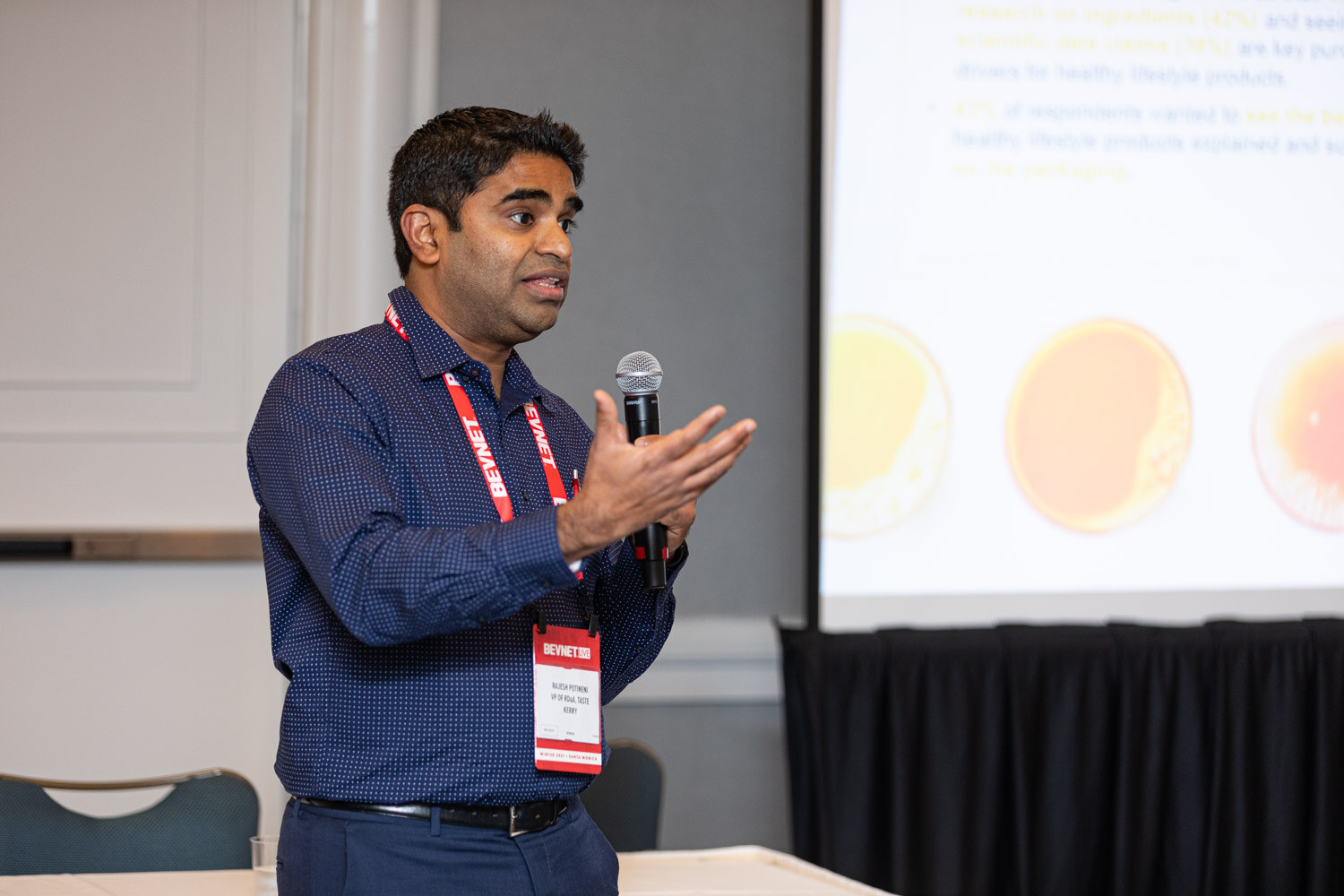 A man in a dark blue shirt with a red BevNET lanyard speaks into a microphone and gestures, with a projected slide behind him.