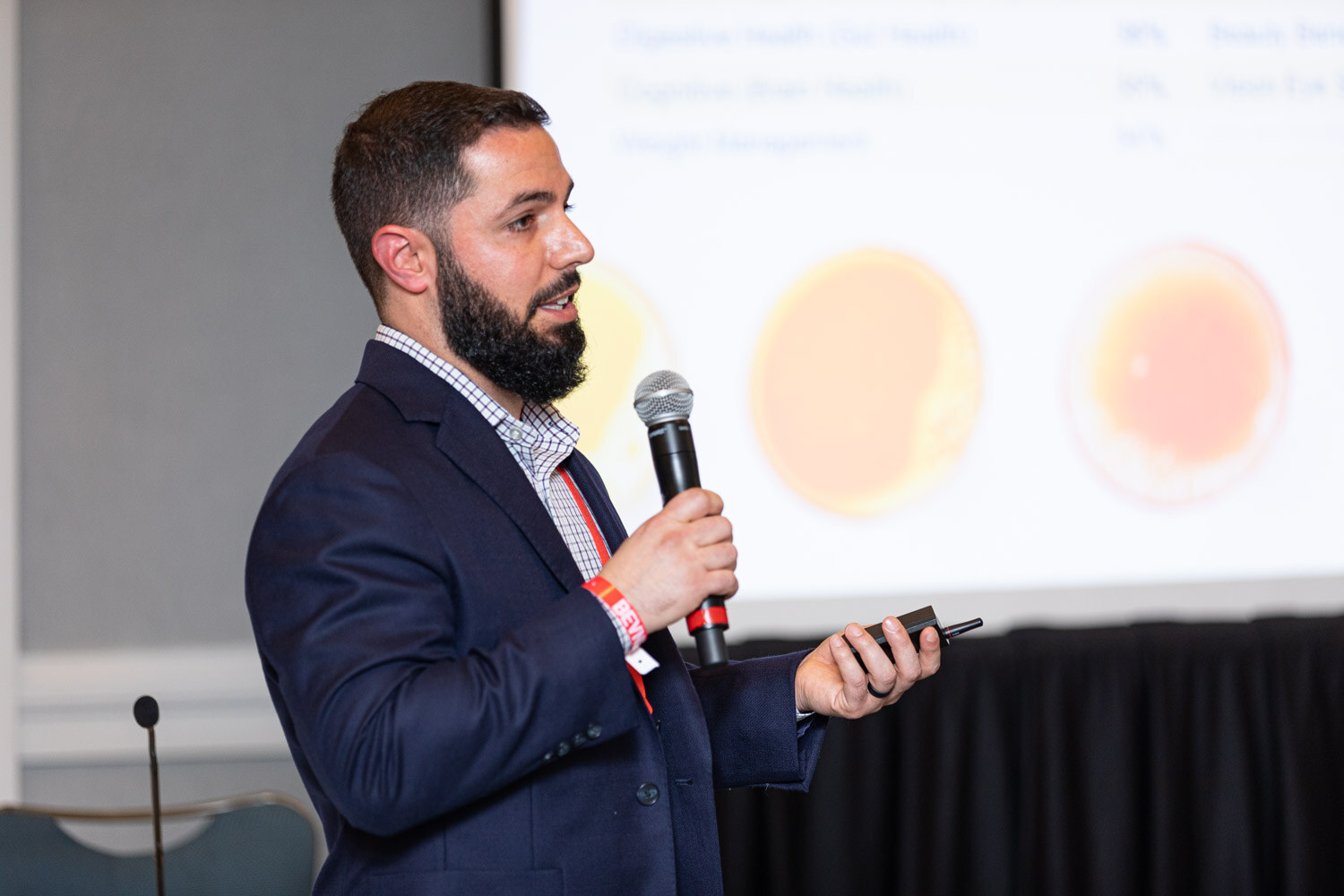 A bearded man in a navy jacket holds a microphone and a clicker in front of a projector screen showing orange circles.