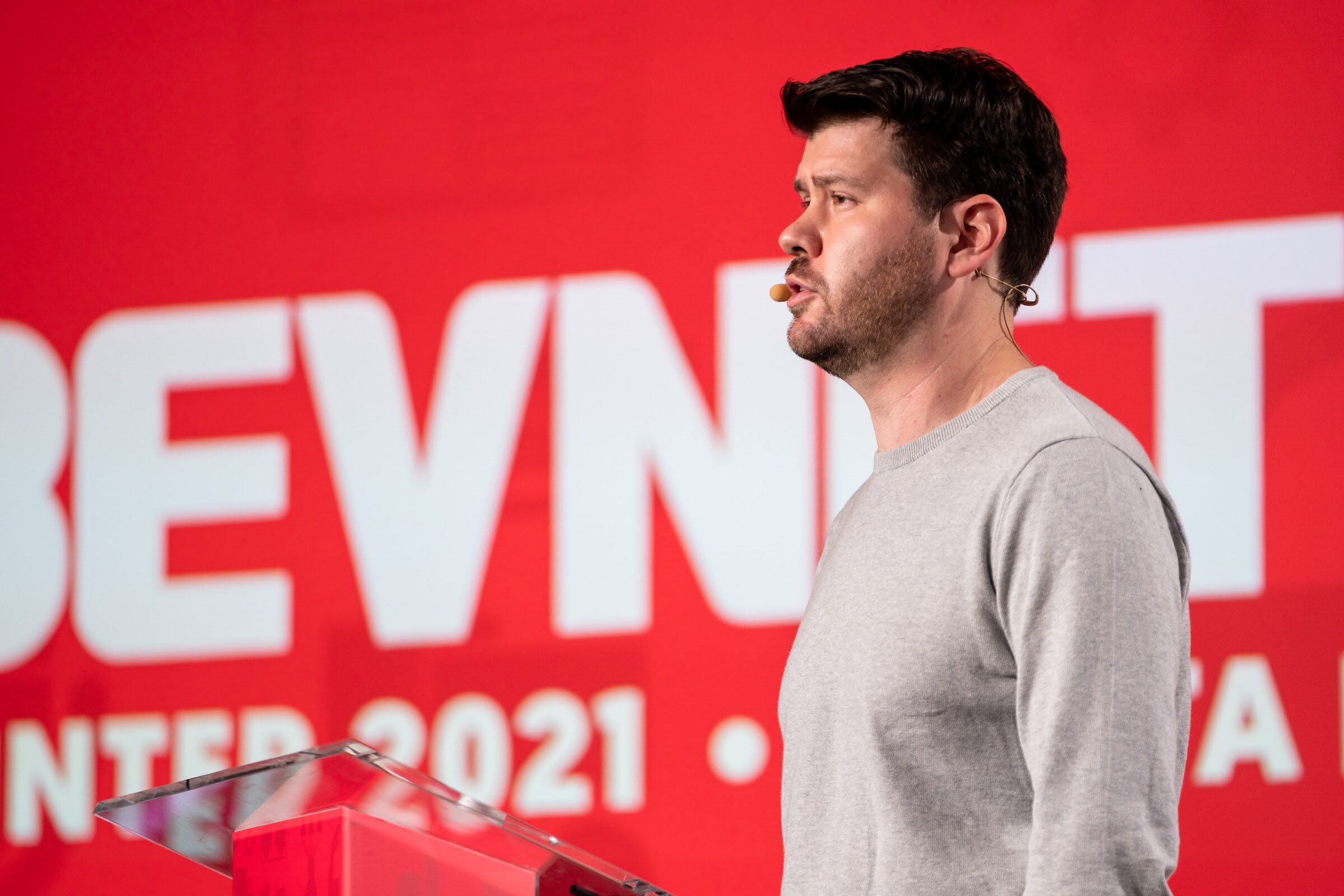 A man in a gray sweater speaks in profile in front of a bright red backdrop with large white BEVNET lettering.