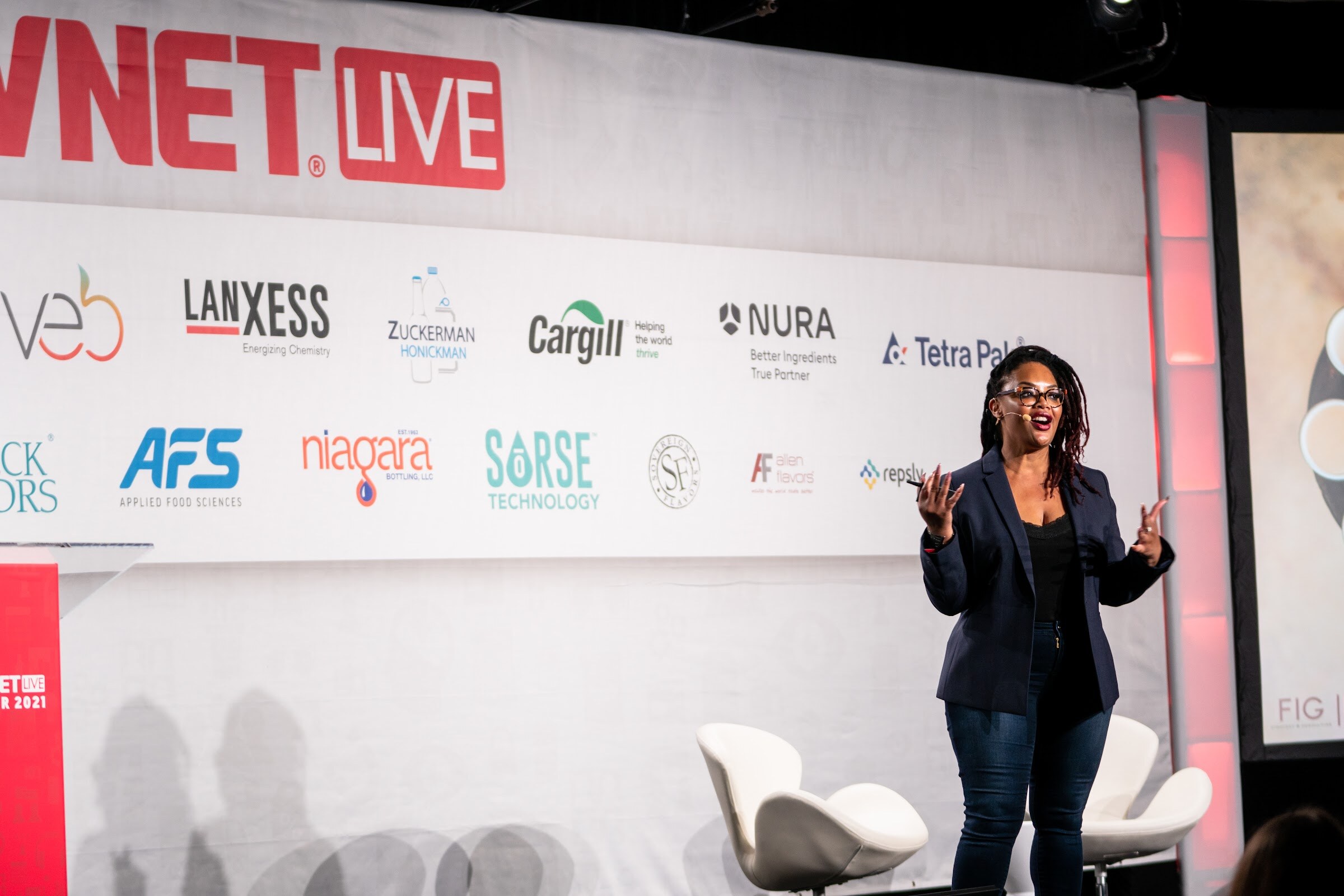 A woman in a navy blazer and jeans stands on a stage with her arms out, in front of a BevNET Live banner and sponsor logos.