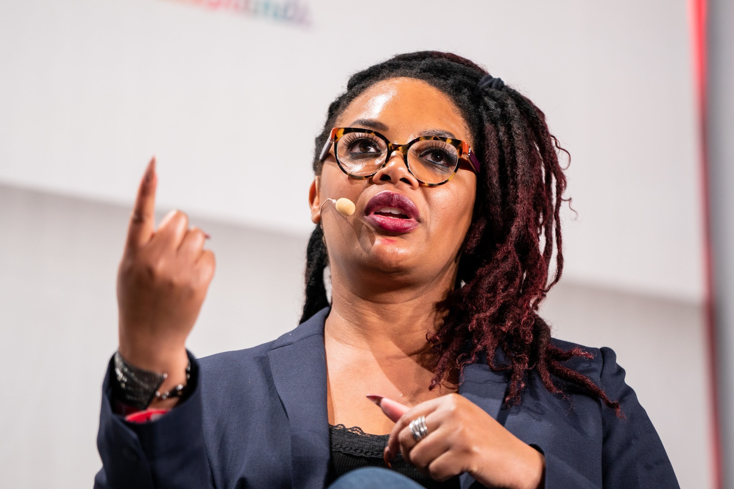 A woman with glasses and braided hair and a headset microphone raises one finger while speaking.