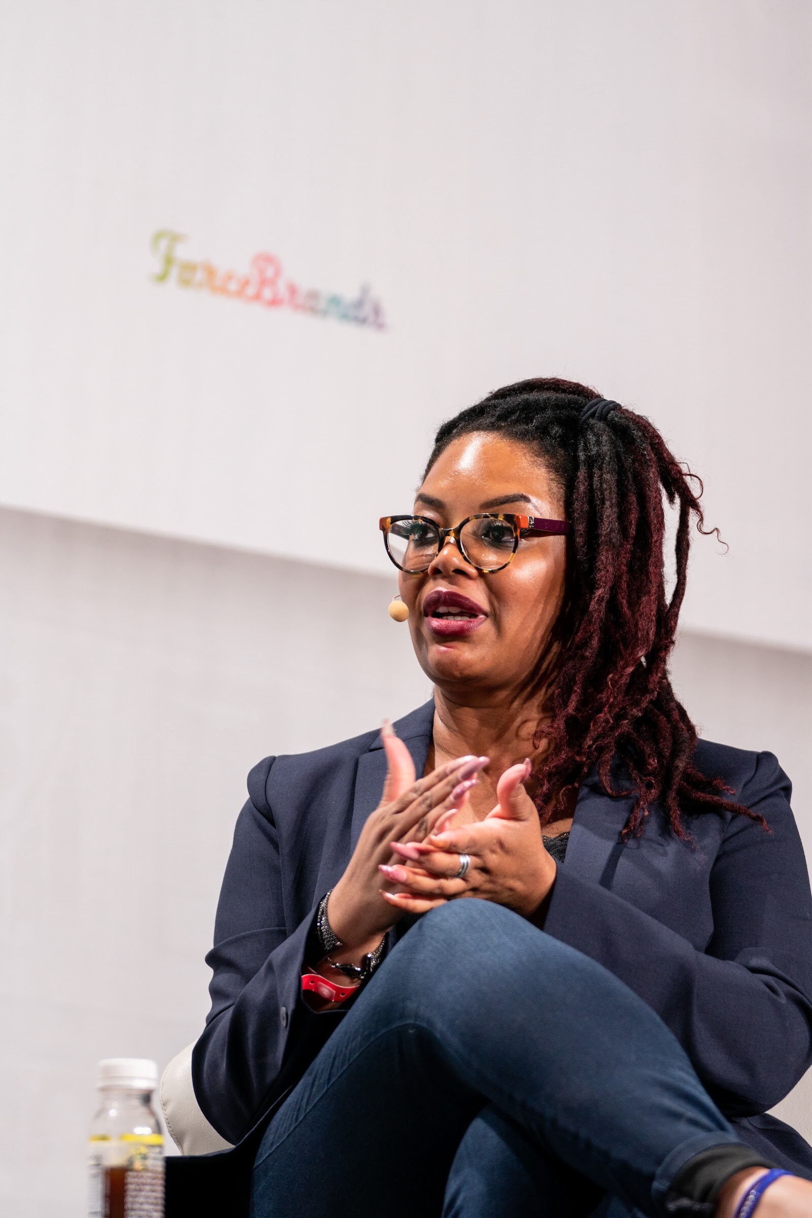 A woman in a navy blazer and glasses with a headset microphone sits with her hands clasped in front of a white wall.