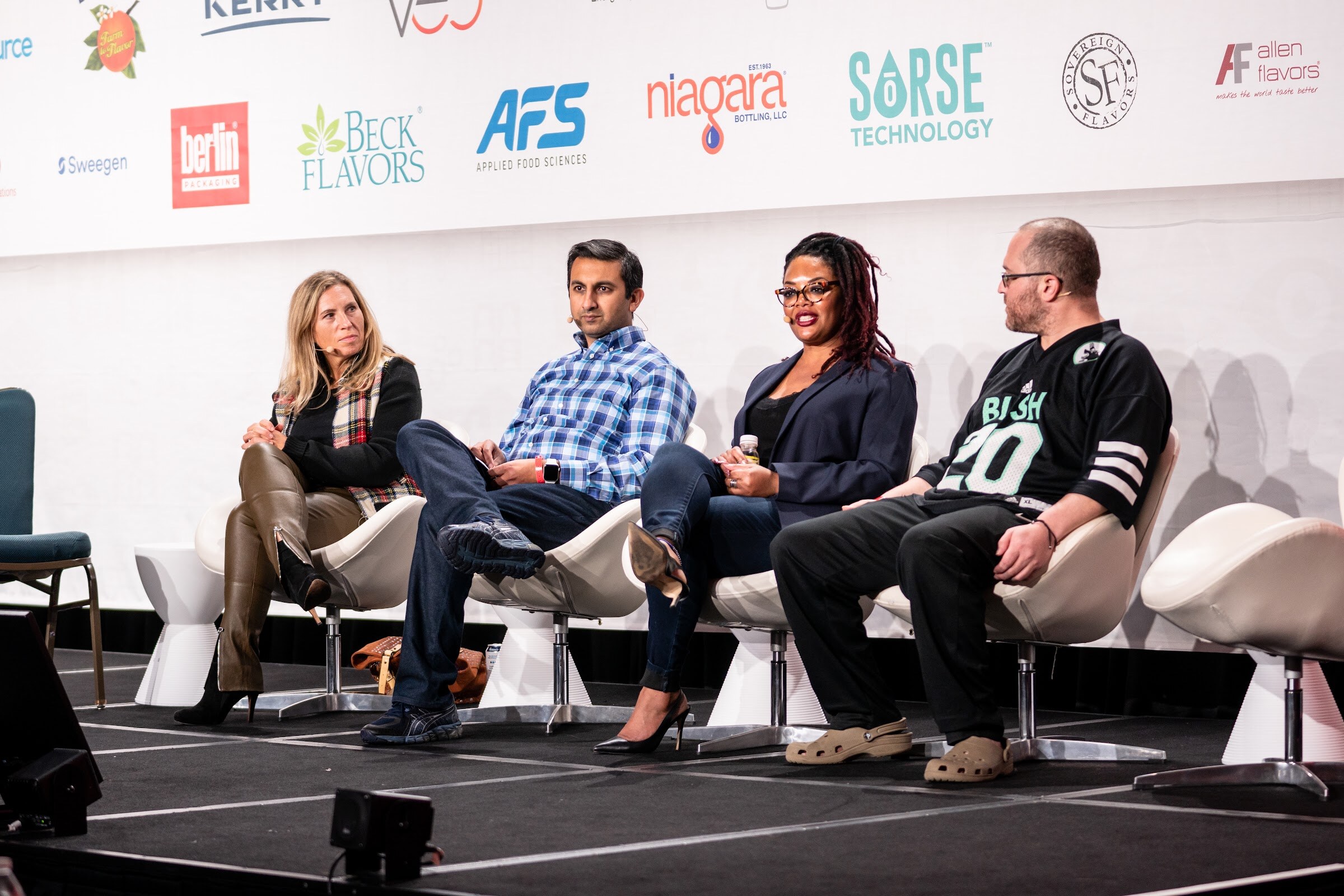 Four people seated in a row on stage in front of a sponsor wall, including a man in a green and black Bush 20 jersey.