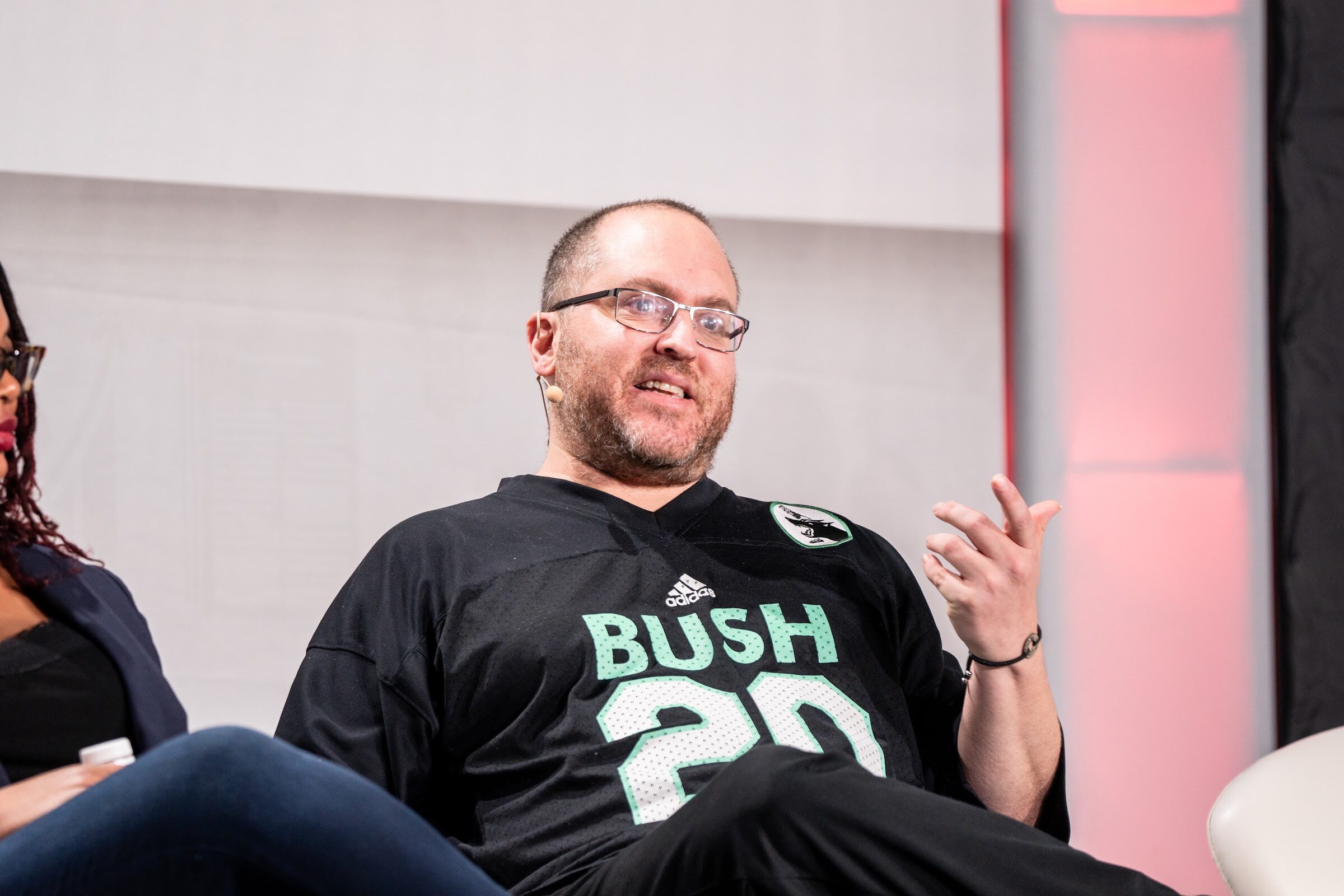 A man with glasses and a beard wearing a black and green Bush 20 jersey gestures with one hand while seated.