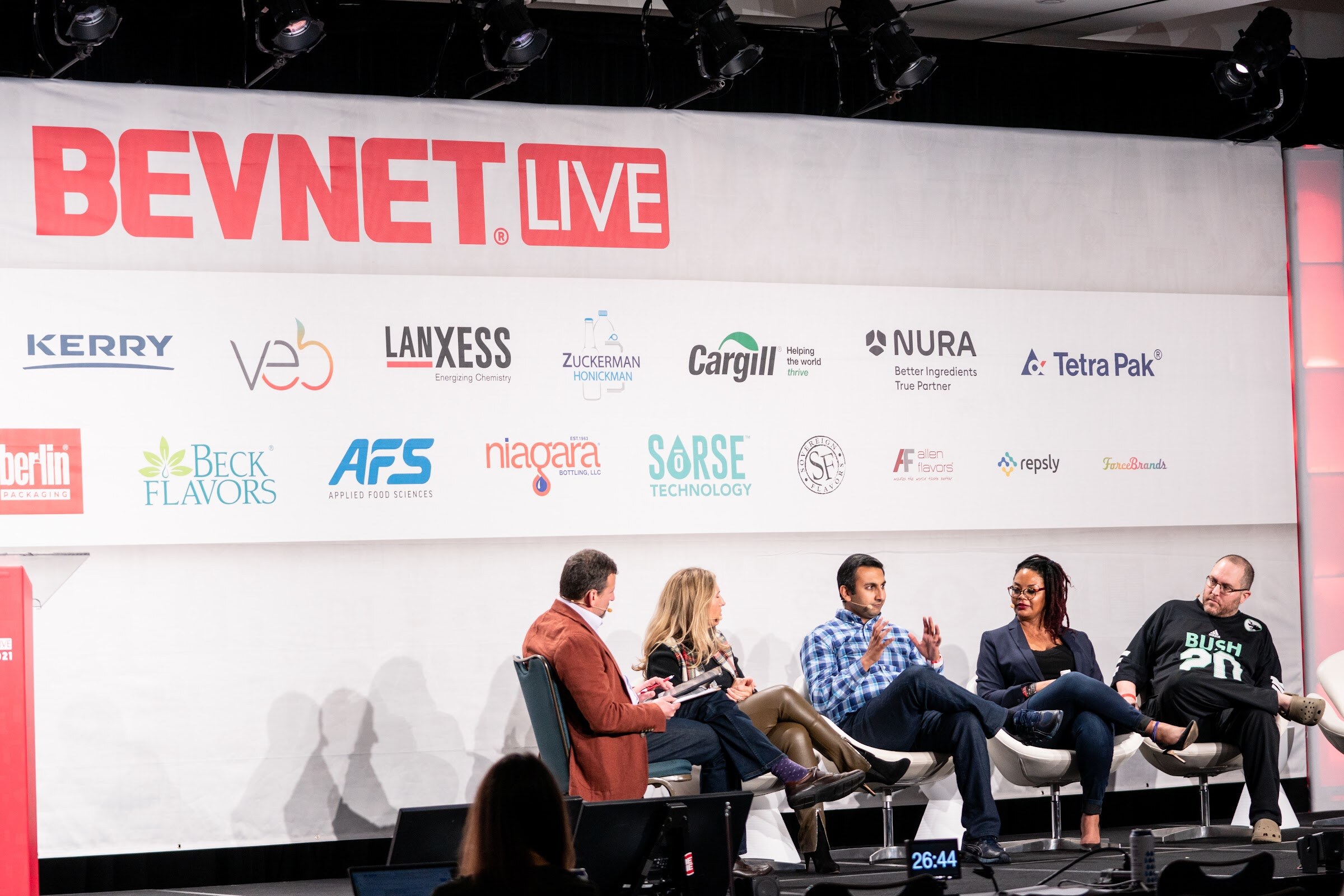 A wide shot of five people seated on a stage below a BevNET Live banner with sponsor logos, with audience members in the foreground.