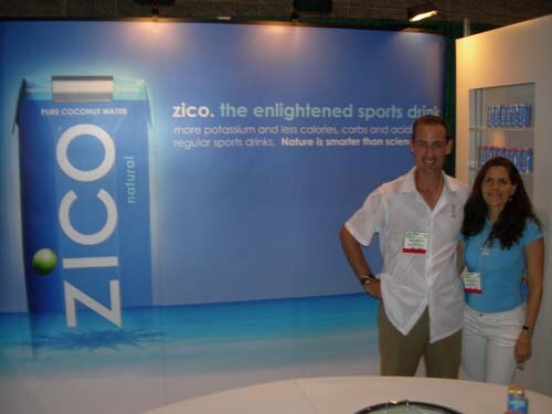A man in a white shirt and a woman in a blue top stand in front of a large blue Zico pure coconut water backdrop with a carton graphic and product shelves.