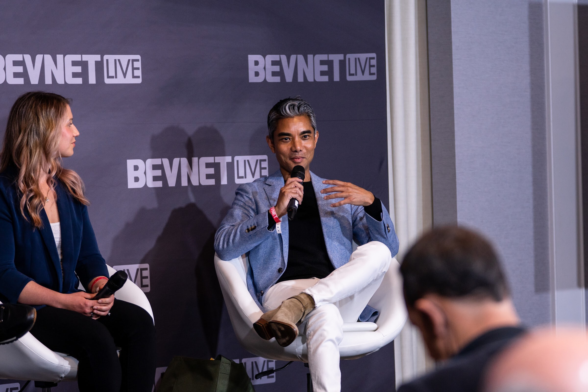 A woman in a navy blazer and a man in a light blue jacket and white pants sit in white chairs in front of a purple BevNET Live wall.