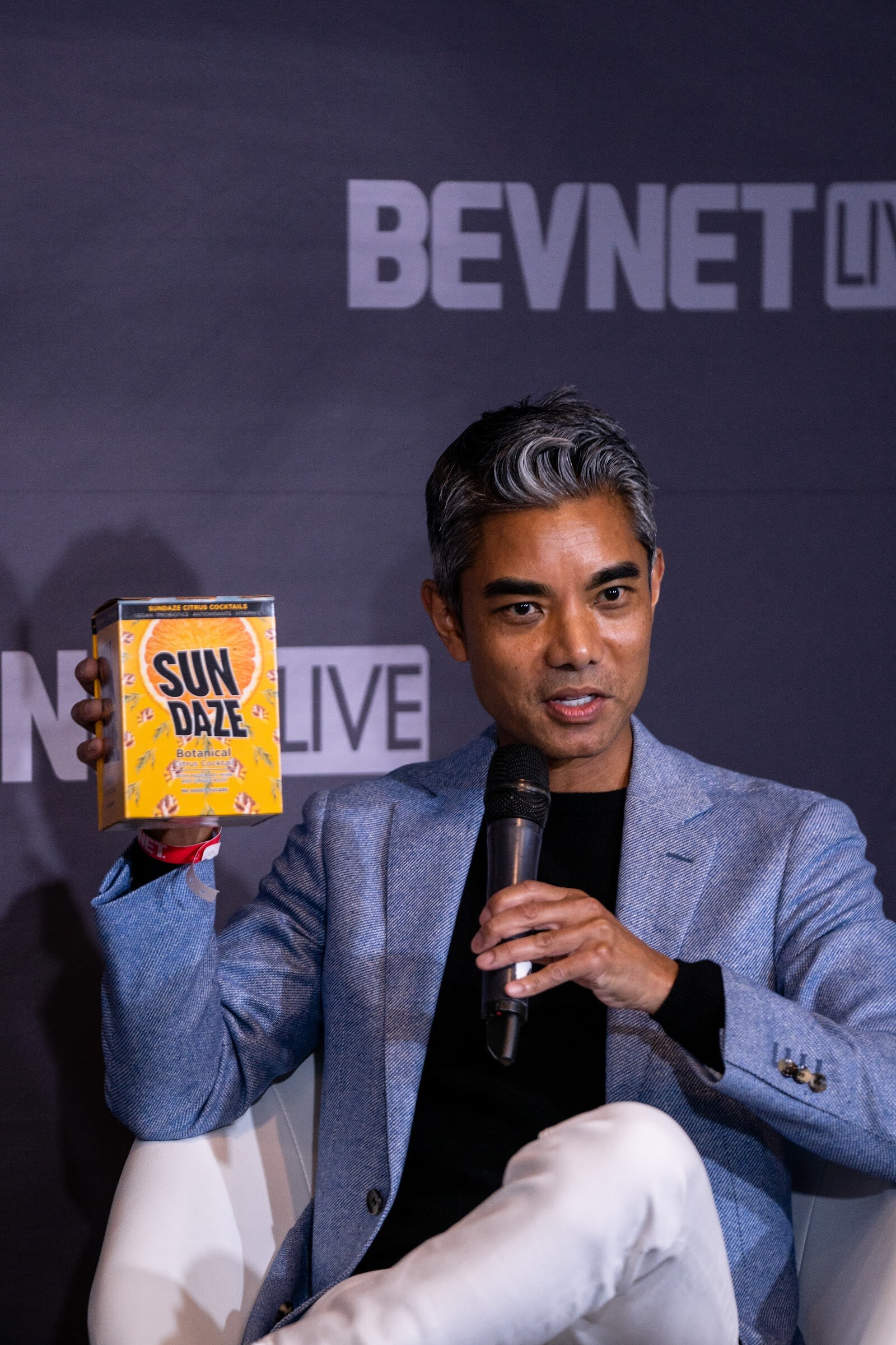A man with gray hair in a light blue blazer holds a yellow box labeled Sun Daze in one hand and a microphone in the other.