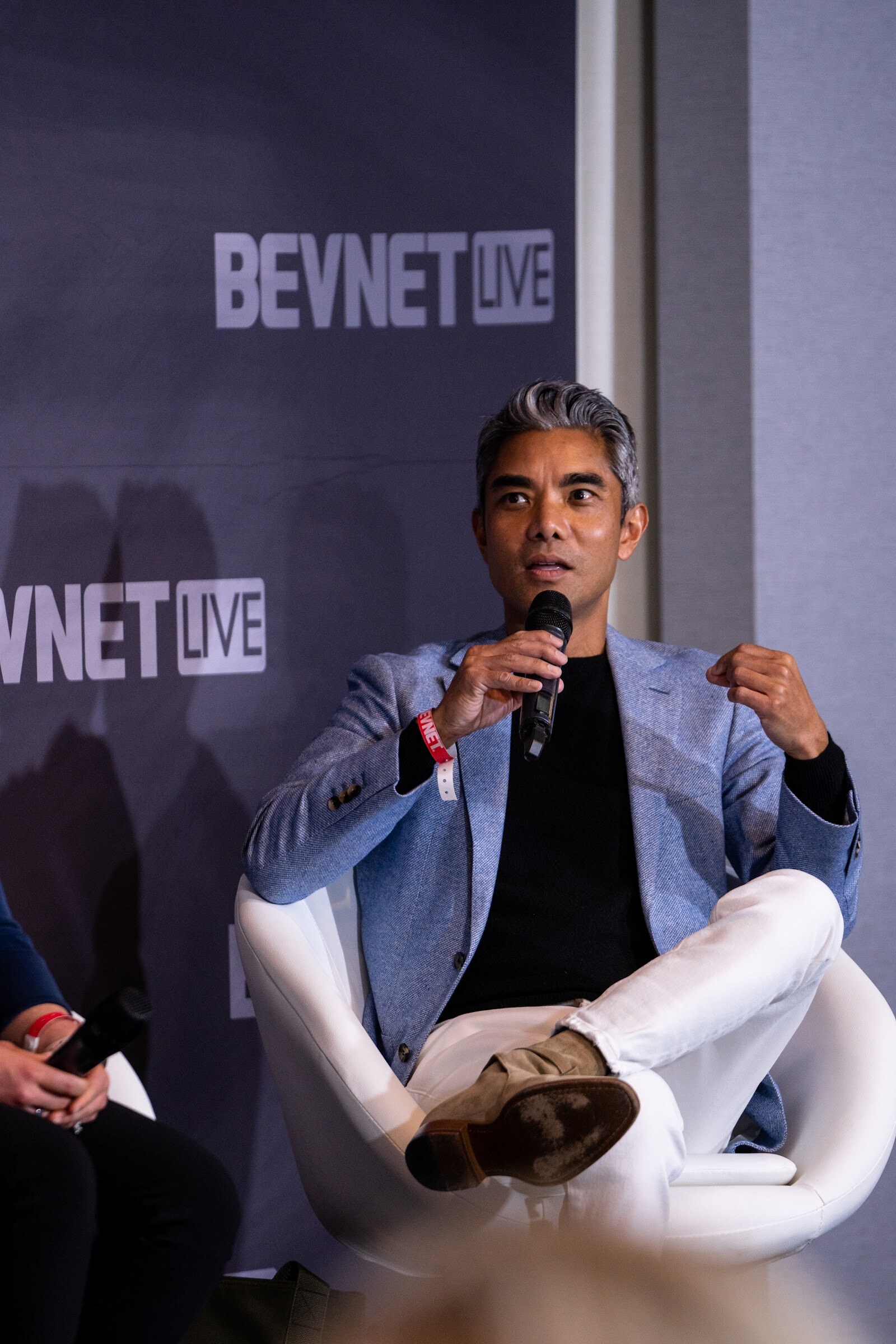 A man with gray hair in a light blue blazer and white pants sits in a white chair holding a microphone in front of a BevNET Live backdrop.