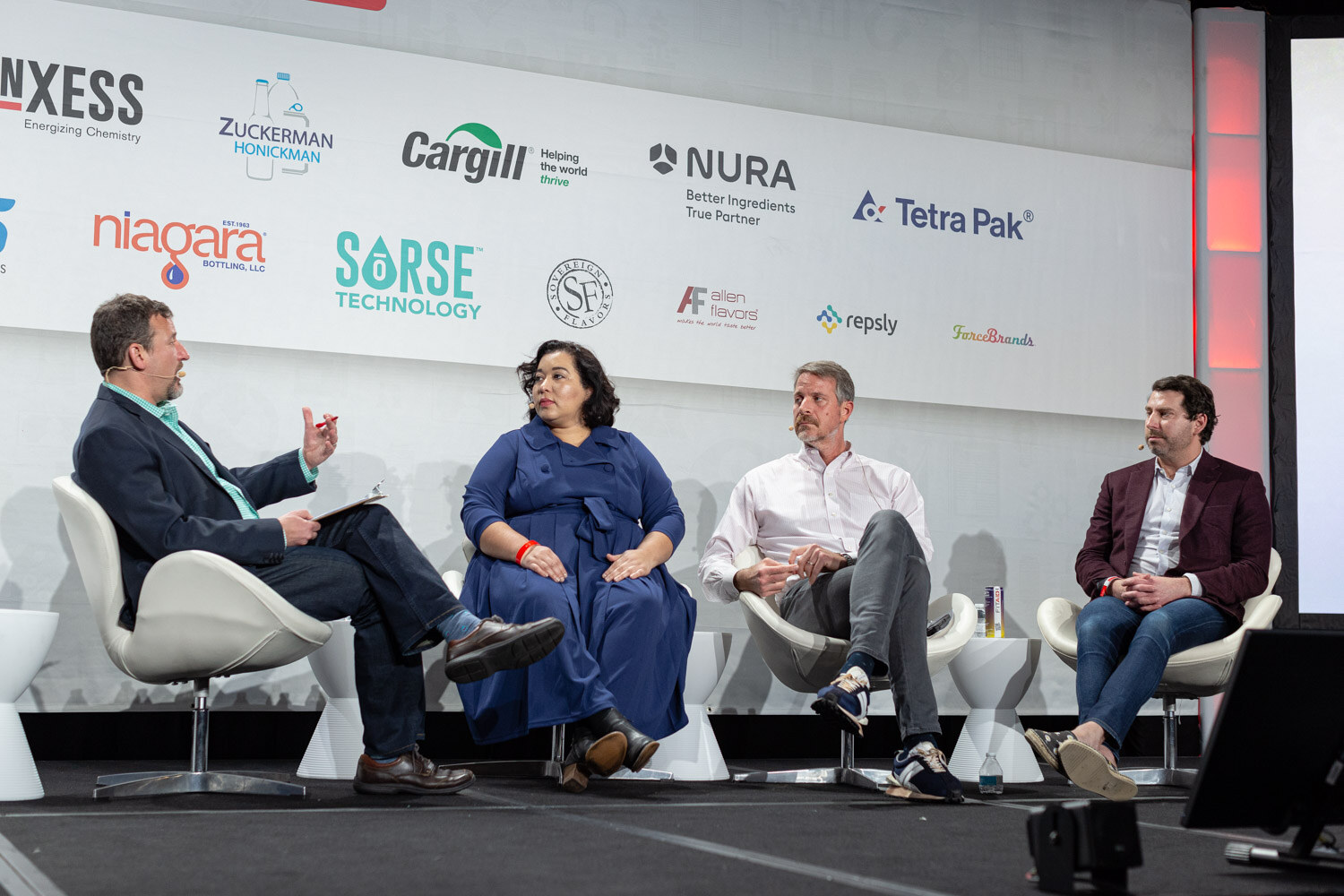 Four people seated in white chairs on stage in front of a sponsor wall with Lanxess, Zuckerman Honickman, Cargill, Nura and Tetra Pak logos.