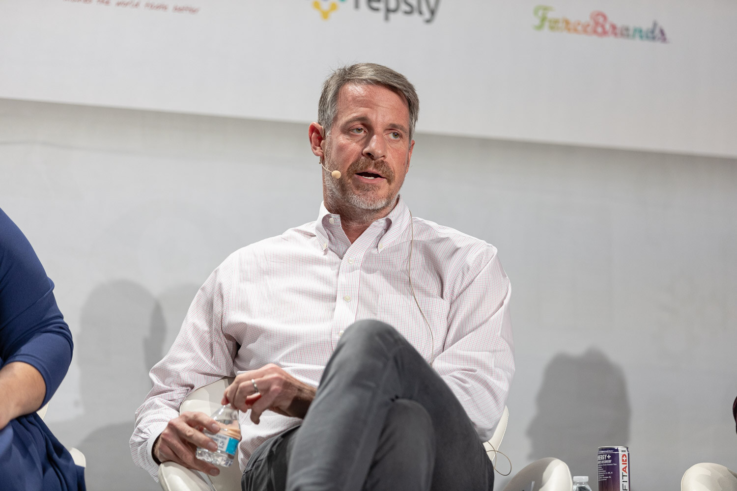 A man with graying hair in a white striped shirt and gray pants sits holding a water bottle and speaks, with a headset microphone.